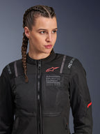 Alpinestars Stella ST-2 Air Jacket, dames motorjas in zwart, voorzien van mesh-panelen voor luchtstroom, 'Alpinestars Exploration Equipment' branding met coördinaten, rood logo-accent op de borst, model met gevlochten haar