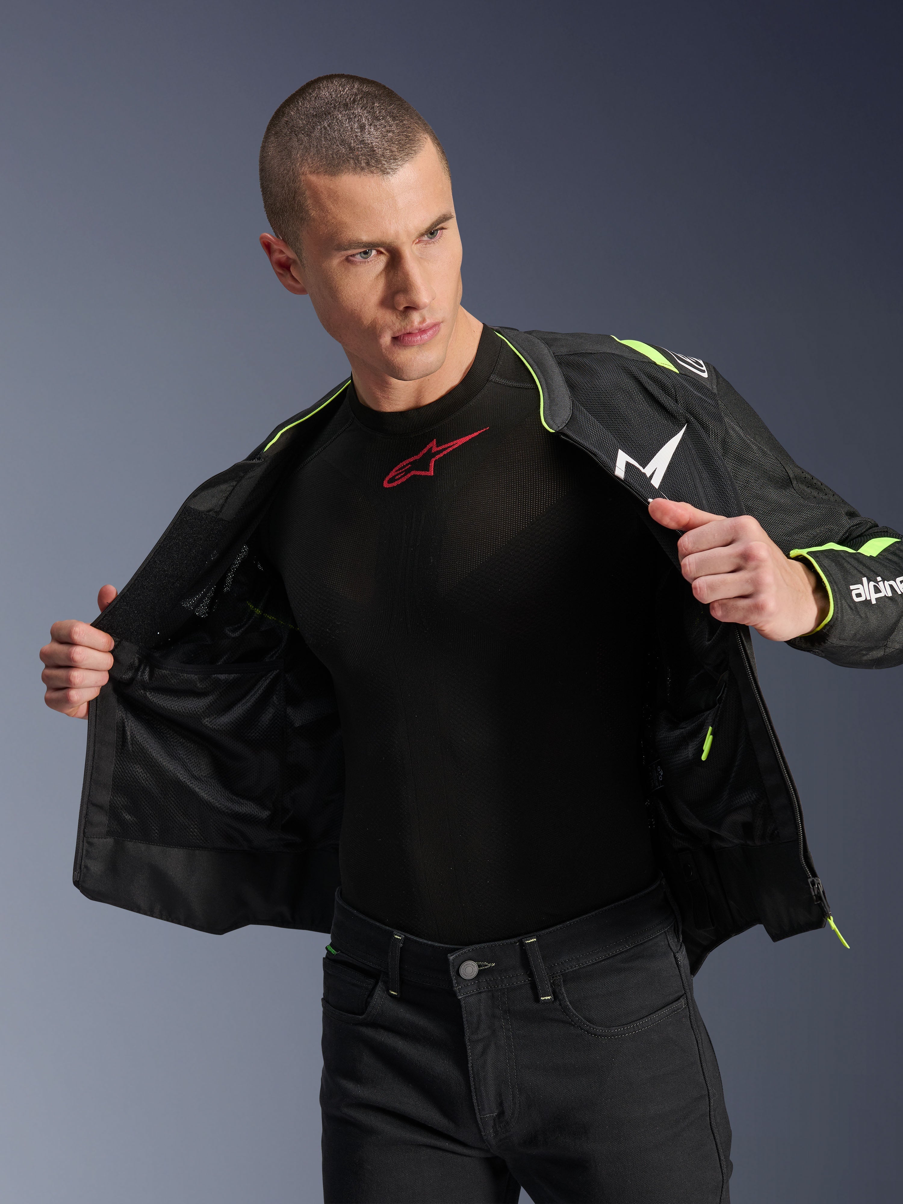 Mannelijk model dat de Alpinestars T-Sps Air V2 Jas in Black Yellow Fluo draagt, open getoond om de interne mesh-constructie en zakken te benadrukken, gecombineerd met een zwarte Alpinestars base layer en zwarte broek, stedelijke motorrijstijl.