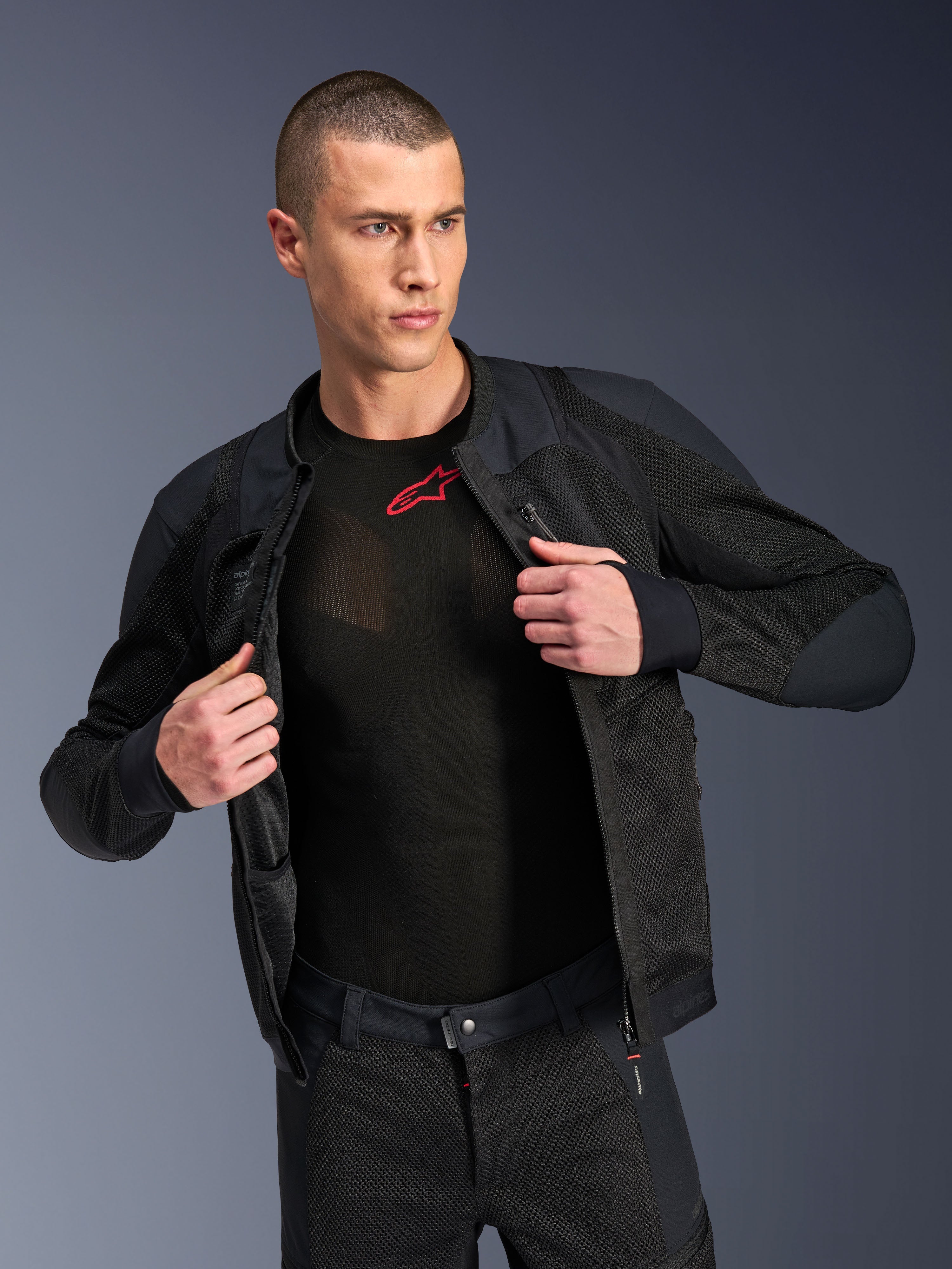 Alpinestars Troop-Air Pro Jas, zwart, hoogwaardige technische gepantserde doorwaaijas met mesh-constructie, open gedragen over een zwarte basislaag met een rood logo, gecombineerd met een bijpassende mesh motorbroek, urban contemporary rijstijl