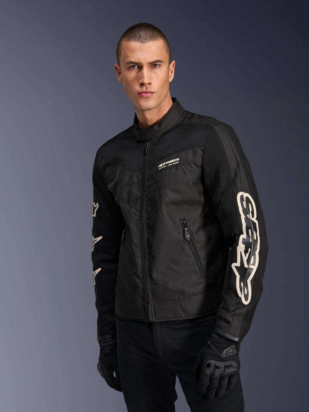 Alpinestars T-Dyno Air Jas in het zwart, mannelijk model draagt een vintage-geïnspireerde textiele motorjas met meshpanelen voor ventilatie, groot wit Alpinestars-logo op de mouwen, gecombineerd met zwarte motorhandschoenen, urban motoruitrusting ontwerp