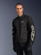 Alpinestars T-Dyno Air Jas in het zwart, mannelijk model draagt een vintage-geïnspireerde textiele motorjas met meshpanelen voor ventilatie, groot wit Alpinestars-logo op de mouwen, gecombineerd met zwarte motorhandschoenen, urban motoruitrusting ontwerp