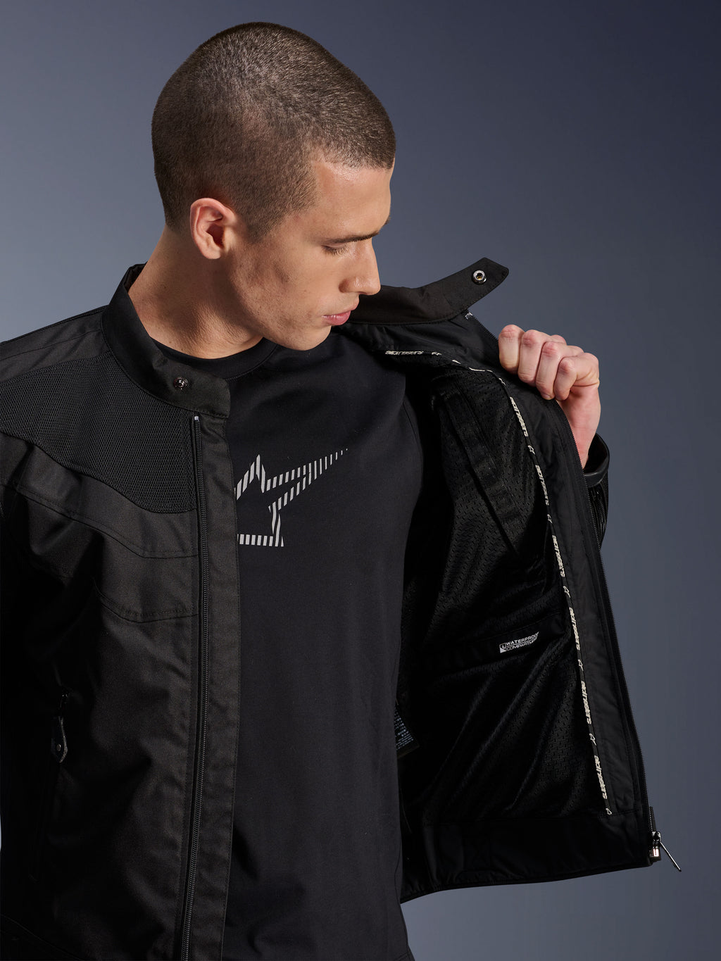 Alpinestars T-Dyno Air Jas, zwart, mannelijk model toont de interne mesh voering en het label van het waterdichte compartiment, gedragen over een zwart grafisch t-shirt, moderne stedelijke motoruitrusting