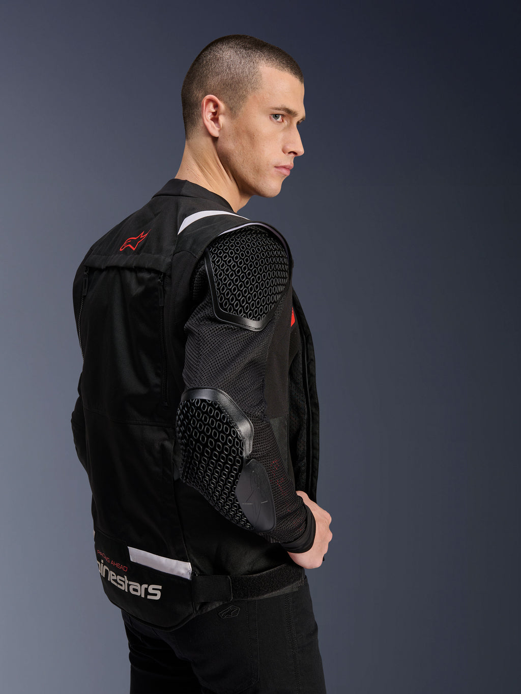 Alpinestars T-Stunt Air Jacket, motorjas, zwart, zijprofiel van een model in de sportief gesneden jas met mesh mouwen en zichtbare honingraatstructuur protectie op de schouder en elleboog, rood logo op de rug, witte branding bij de taille.