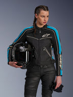 Vrouw draagt Alpinestars Stella T-Dyno Air jas in Black Light Blue Ecru, houdt een zwarte carbon motorhelm en handschoenen vast, voorzien van een getailleerde pasvorm met 3D-meshpanelen en vintage-geïnspireerd ontwerp, eigentijdse motorkleding