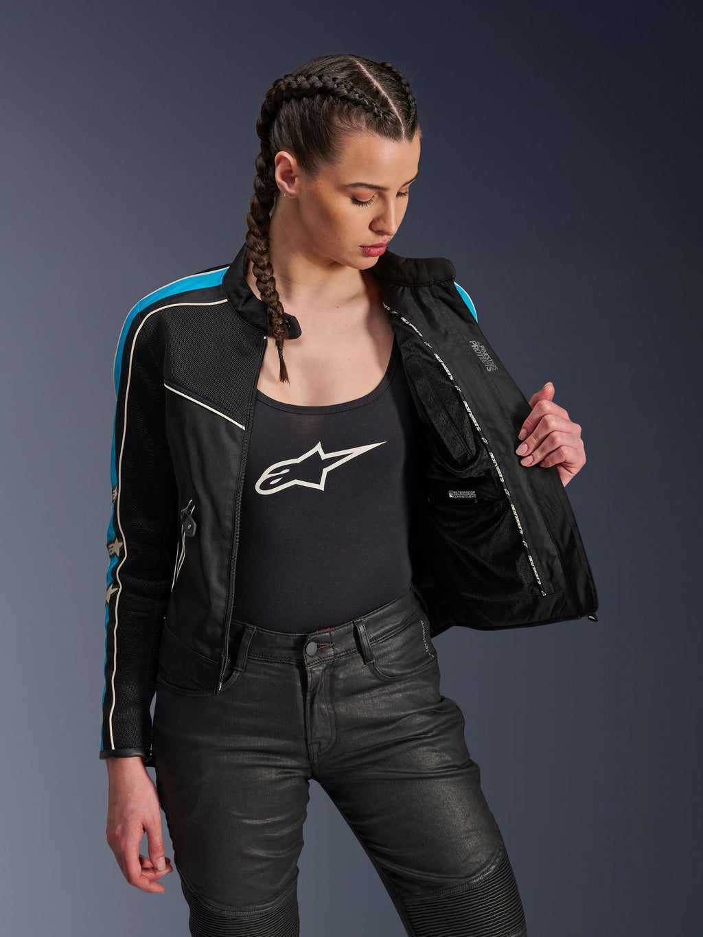 Vrouw die de Alpinestars Stella T-Dyno Air Jas in Black Light Blue Ecru draagt en de jas openhoudt om de binnenste mesh voering en de waterdichte zak te tonen, gecombineerd met een zwarte tanktop met logo en een zwarte motorbroek, vintage-geïnspireerde urban motorkleding.