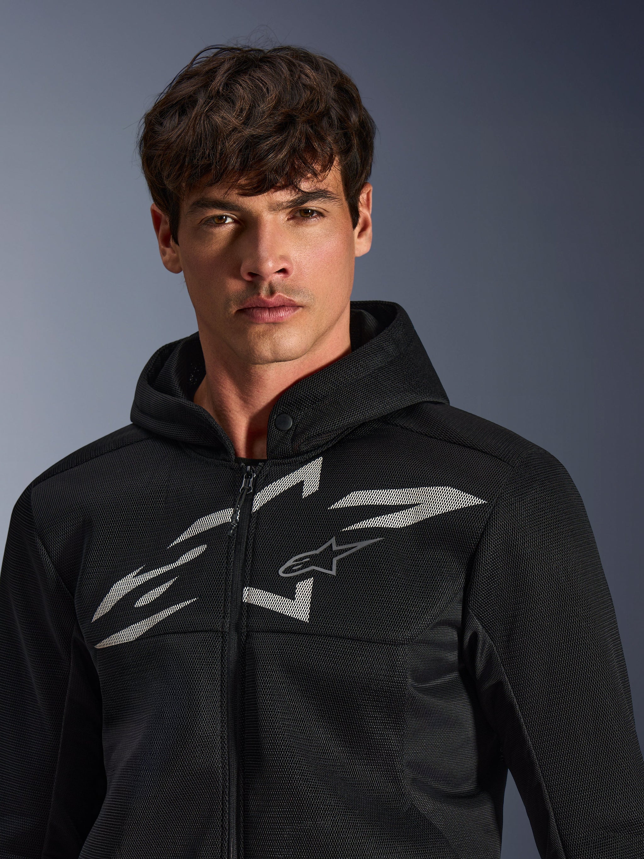 Alpinestars Chrome Superairflow Hoodie, zwarte motorhoodie, voorzien van 3D-meshconstructie met een groot wit grafisch logo op de borst, gedragen door een model, urban sport-rijuitrusting met een modern geventileerd ontwerp
