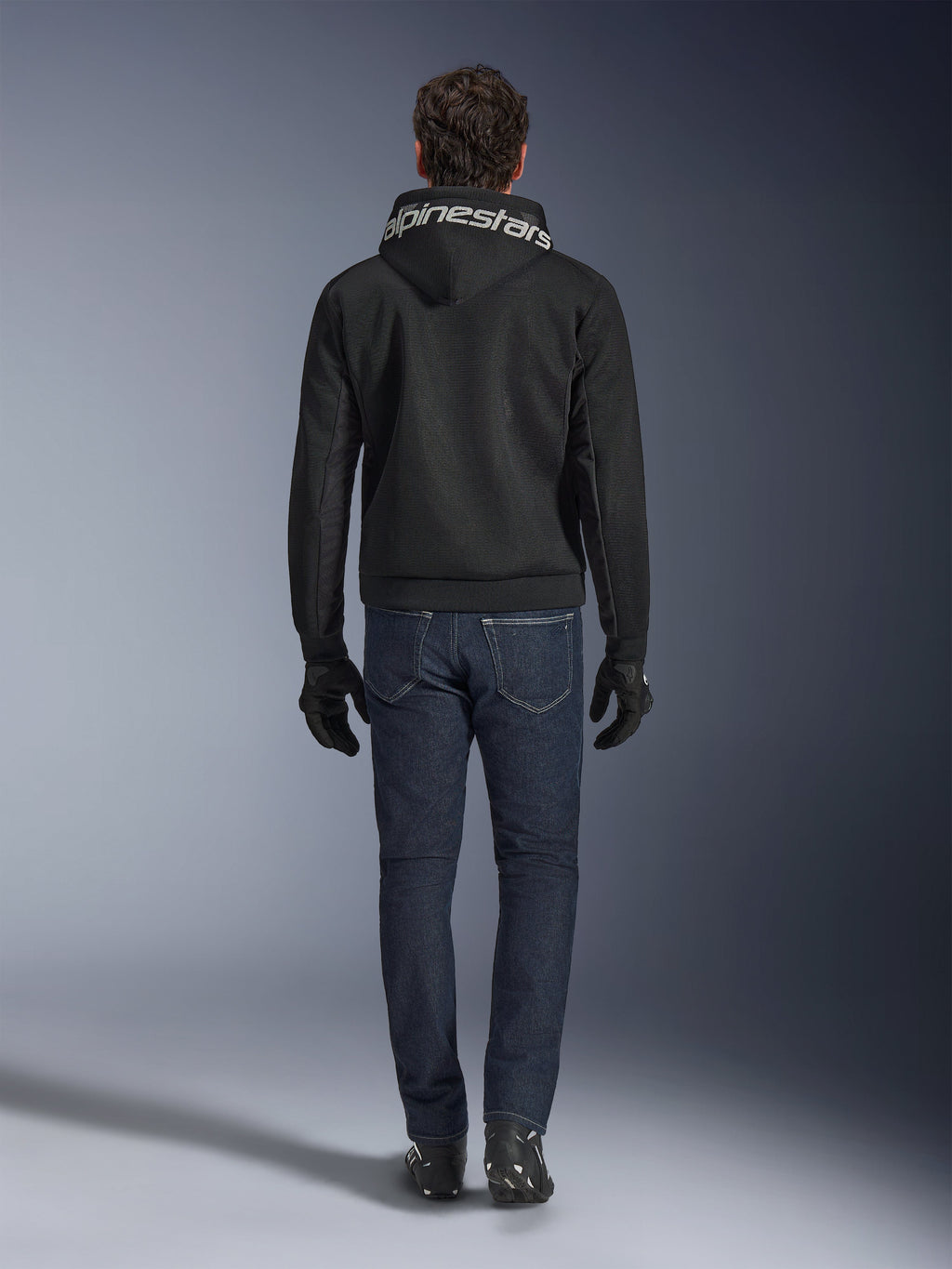 Alpinestars Chrome Superairflow Hoodie, zwarte motorhoodie met witte branding op de capuchon, achteraanzicht, gedragen met een donkerblauwe jeans, zwarte handschoenen en zwart-witte laarzen, urban sportieve motoruitrusting