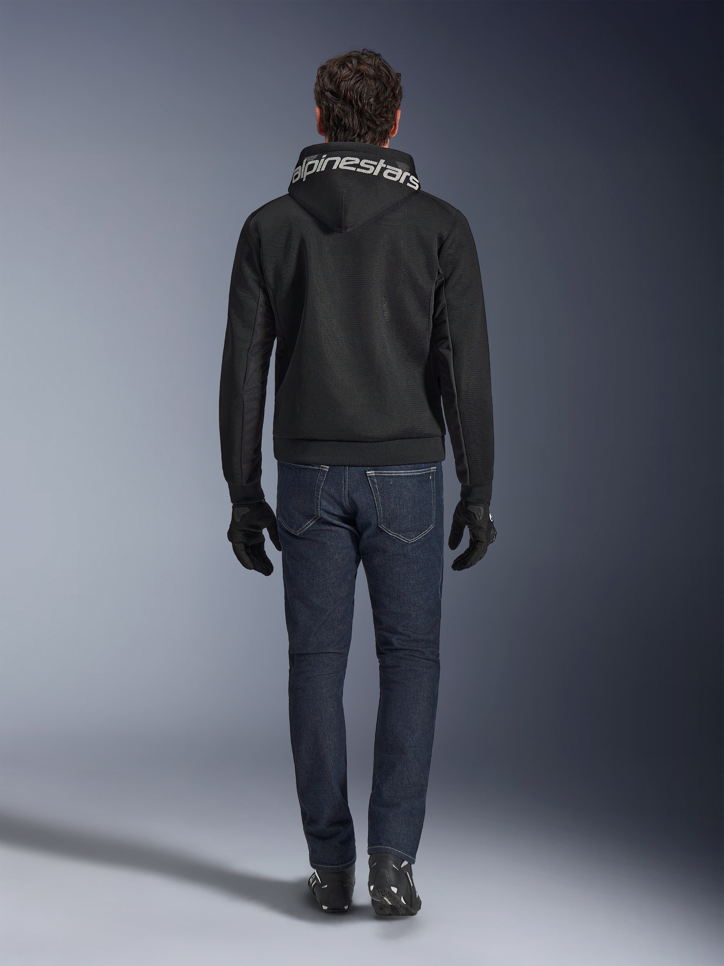 Alpinestars Chrome Superairflow Hoodie, zwarte motorhoodie met witte branding op de capuchon, achteraanzicht, gedragen met een donkerblauwe jeans, zwarte handschoenen en zwart-witte laarzen, urban sportieve motoruitrusting
