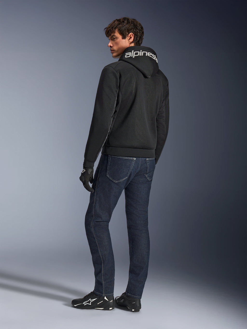 Alpinestars Chrome Superairflow Hoodie, zwart, achteraanzicht op mannelijk model, voorzien van witte Alpinestars-branding op de capuchon, gedragen met een donkere denim jeans en zwarte handschoenen, urban motoruitrusting ontwerp
