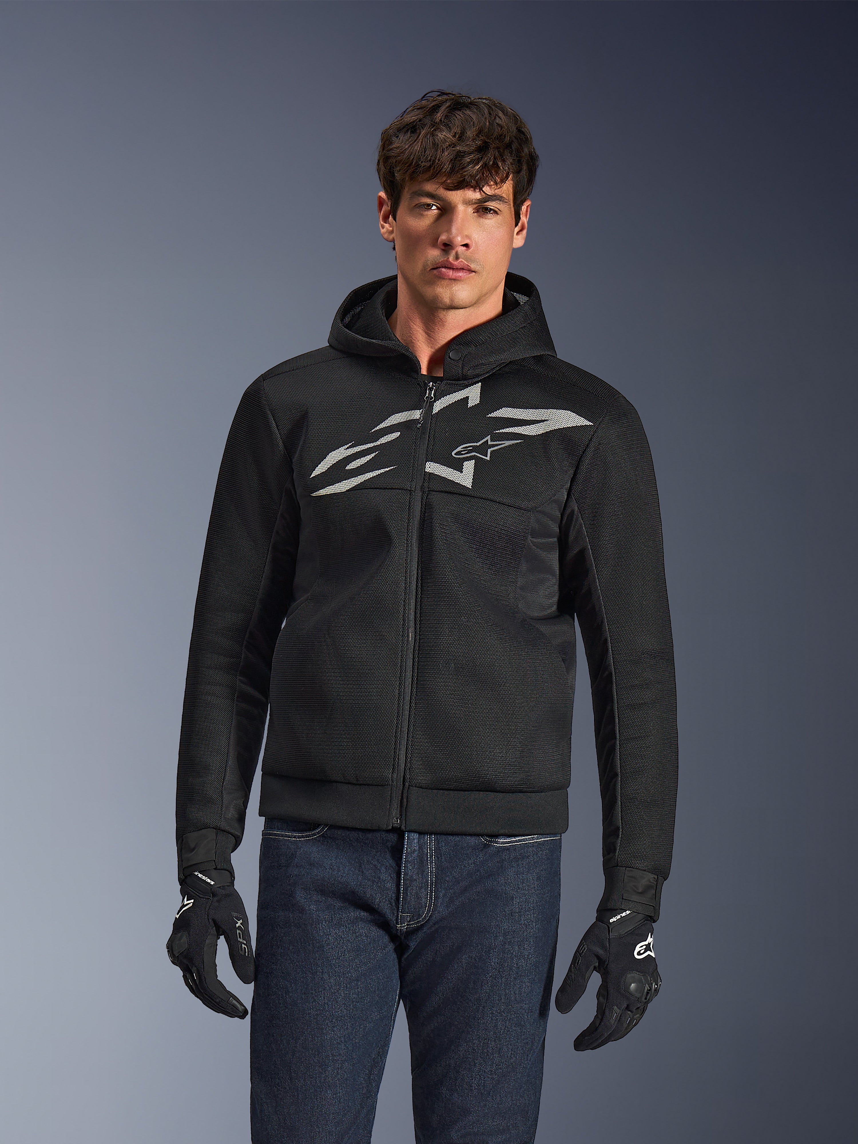 Alpinestars Chrome Superairflow Hoodie, zwarte motorhoodie met ritssluiting en een groot wit logo op de borst, mesh airflow-constructie, gedragen met een blauwe jeans en zwarte handschoenen, sportieve stedelijke motorkleding
