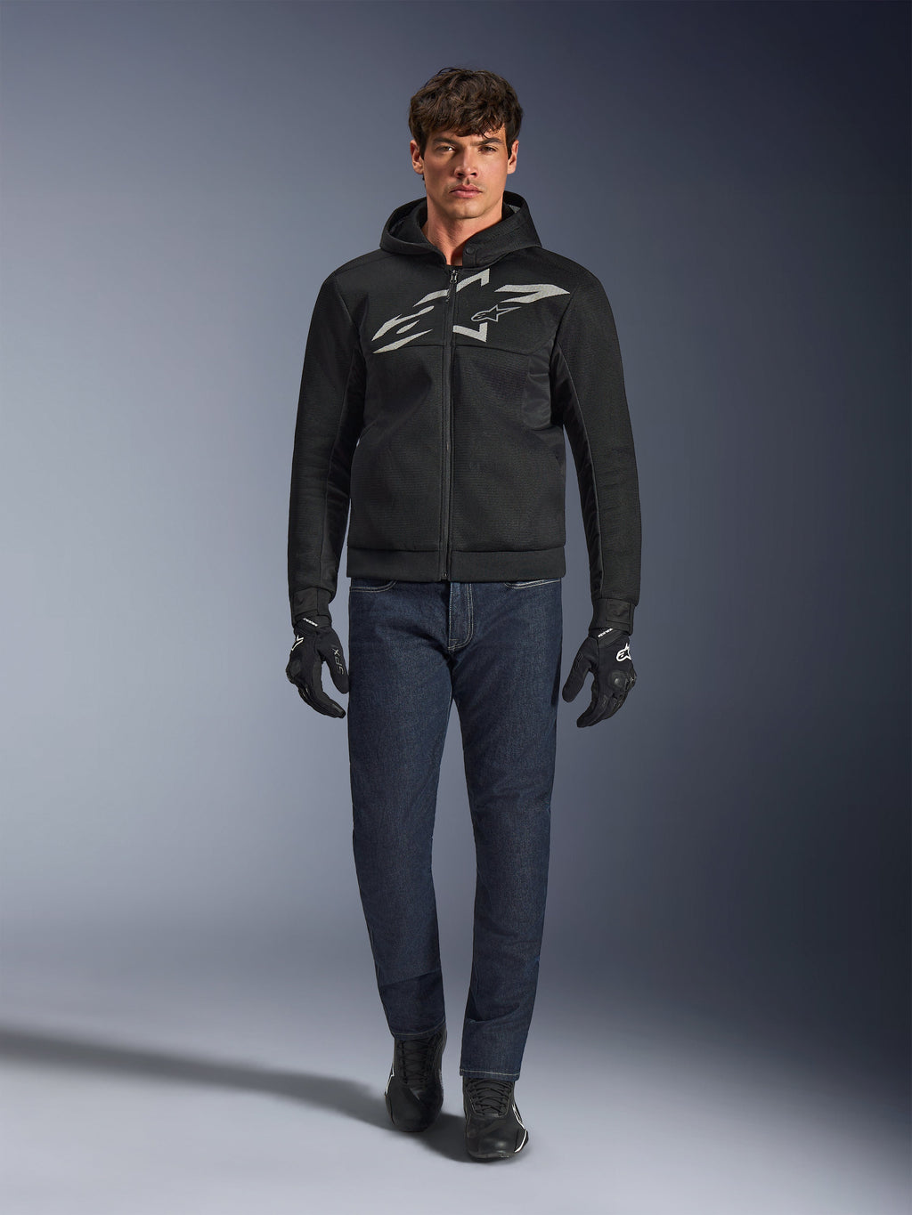 Motorrijder die de Alpinestars Chrome Superairflow Hoodie in zwart en grijs draagt, gecombineerd met een donkerblauwe jeans, zwarte motorhandschoenen en zwarte schoenen. De hoodie is voorzien van een groot gestileerd logo op de borst en een 3D mesh-textuur voor prestaties bij hoge temperaturen.
