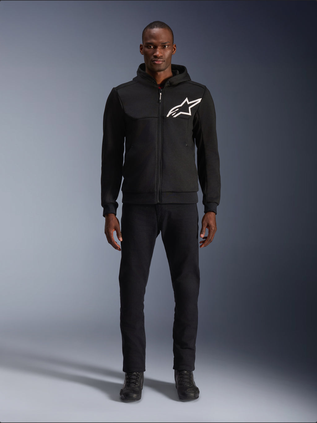 Alpinestars Chrome V2 Sport Hoodie, zwart-witte motorhoodie met een groot wit Alpinestars-logo op de borst, ritssluiting en capuchon, gedragen door een model met zwarte broek en laarzen, urban-stijl beschermende motorkleding.
