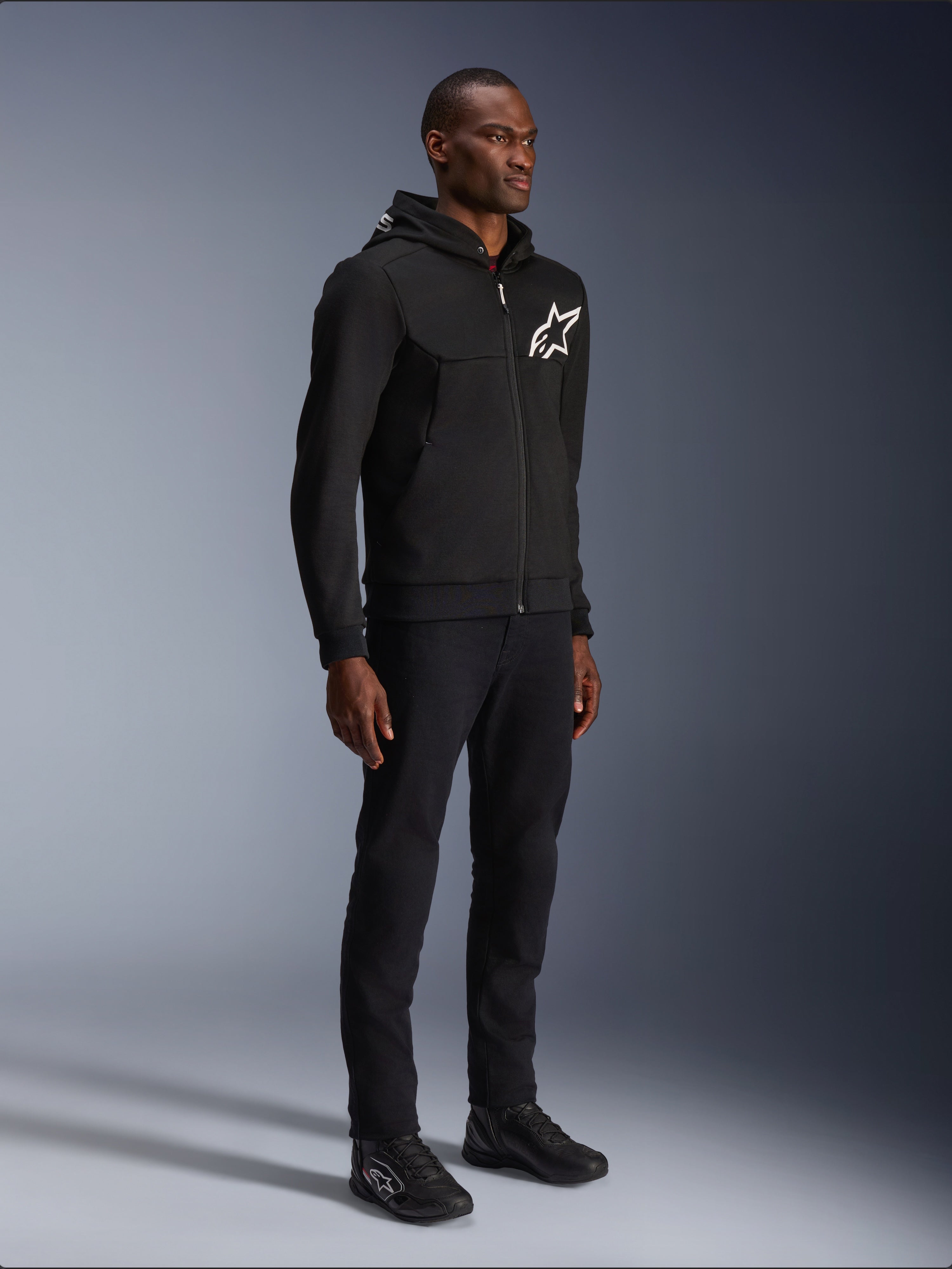 Alpinestars Chrome V2 Sport Hoodie, zwarte motorhoodie met wit sterlogo, gedragen door een mannelijk model met bijpassende zwarte broek en motorsneakers, urban lifestyle motorkleding