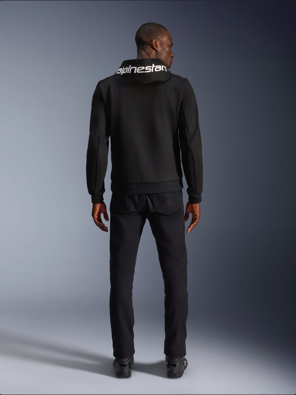 Alpinestars Chrome V2 Sport Hoodie, motorhoodie, zwart, achteraanzicht van model die een zwarte tech fleece hoodie draagt met wit Alpinestars-logo op de capuchon, gecombineerd met zwarte broek en laarzen, casual urban rijstijl