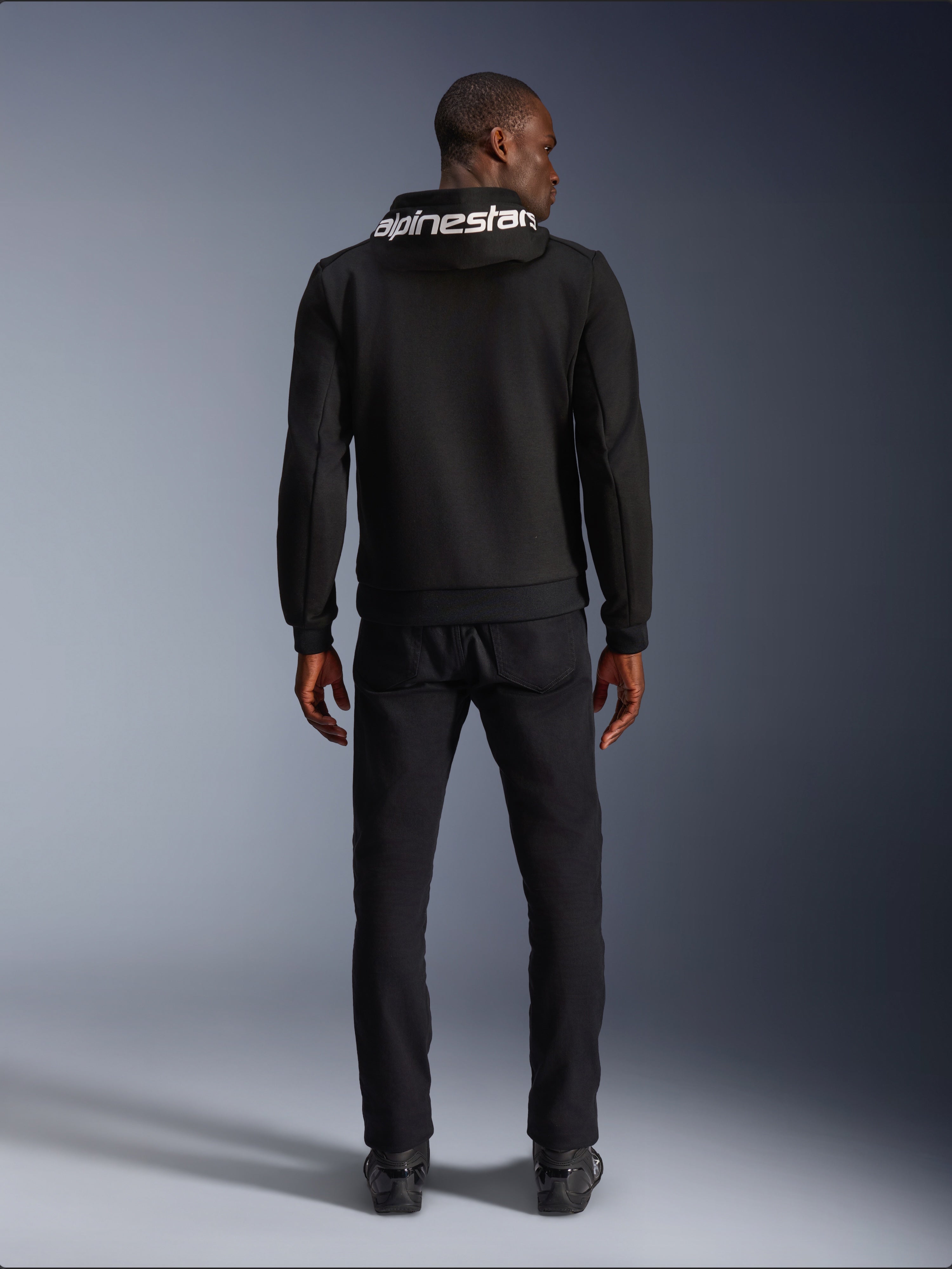 Alpinestars Chrome V2 Sport Hoodie, motorhoodie, zwart, achteraanzicht van model die een zwarte tech fleece hoodie draagt met wit Alpinestars-logo op de capuchon, gecombineerd met zwarte broek en laarzen, casual urban rijstijl