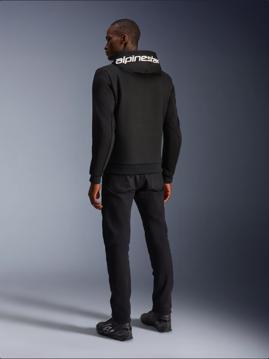 Alpinestars Chrome V2 Sport Hoodie, zwart, achteraanzicht met witte Alpinestars-branding op de capuchon, gedragen met zwarte broek en motorschoenen, casual stedelijke motorkleding met een atletisch silhouet