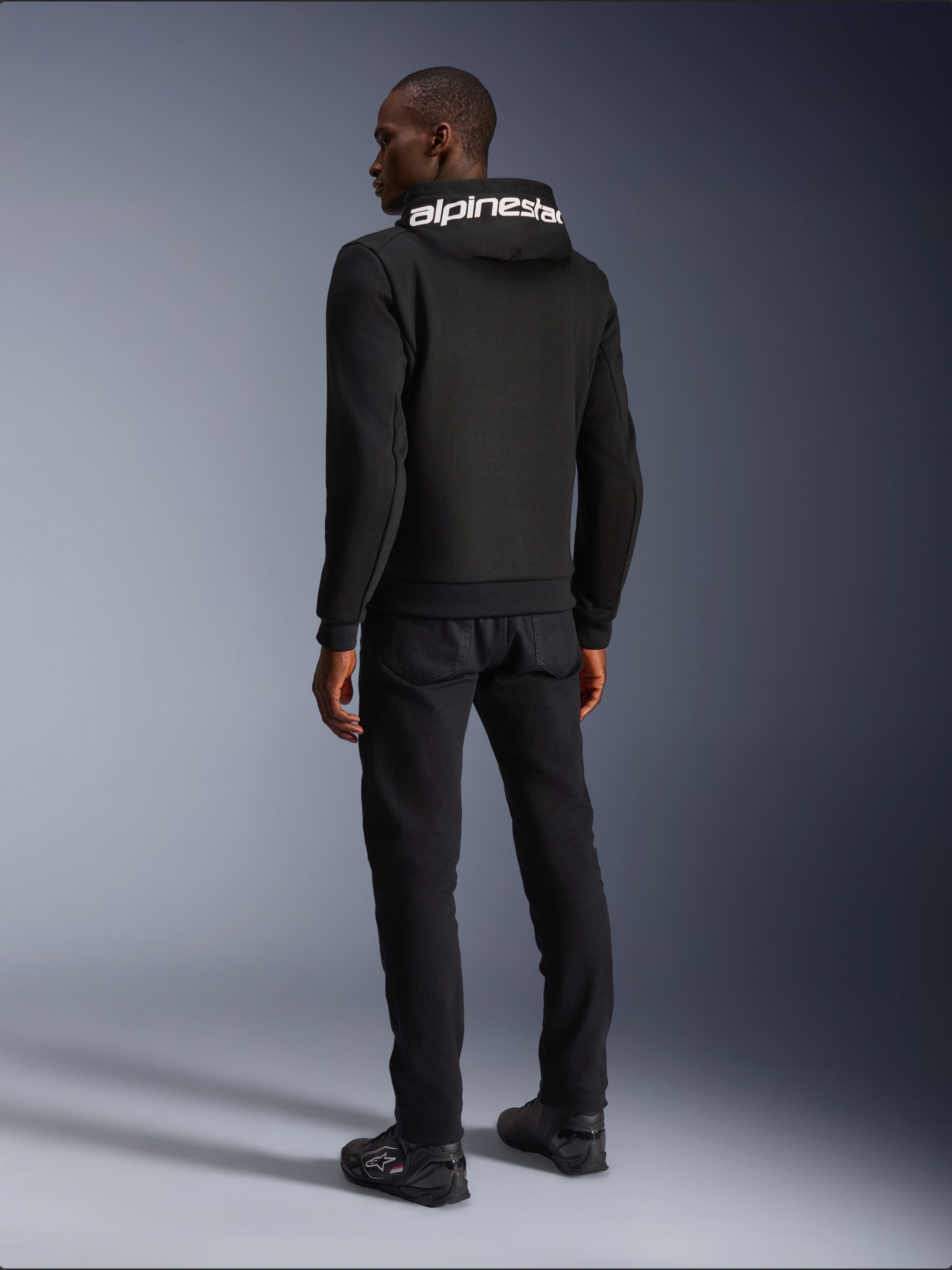 Alpinestars Chrome V2 Sport Hoodie, zwart, achteraanzicht met witte Alpinestars-branding op de capuchon, gedragen met zwarte broek en motorschoenen, casual stedelijke motorkleding met een atletisch silhouet
