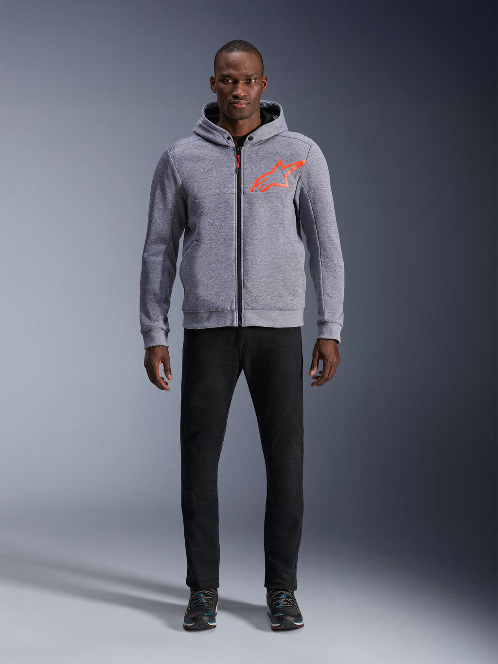 Alpinestars Chrome V2 Sport Hoodie in Gray Heather Red Fluo, lichtgrijze tech fleece hoodie met een fel oranjerood ster-logo op de borst, model draagt zwarte broek en donkere sneakers, casual stedelijke motorstijl