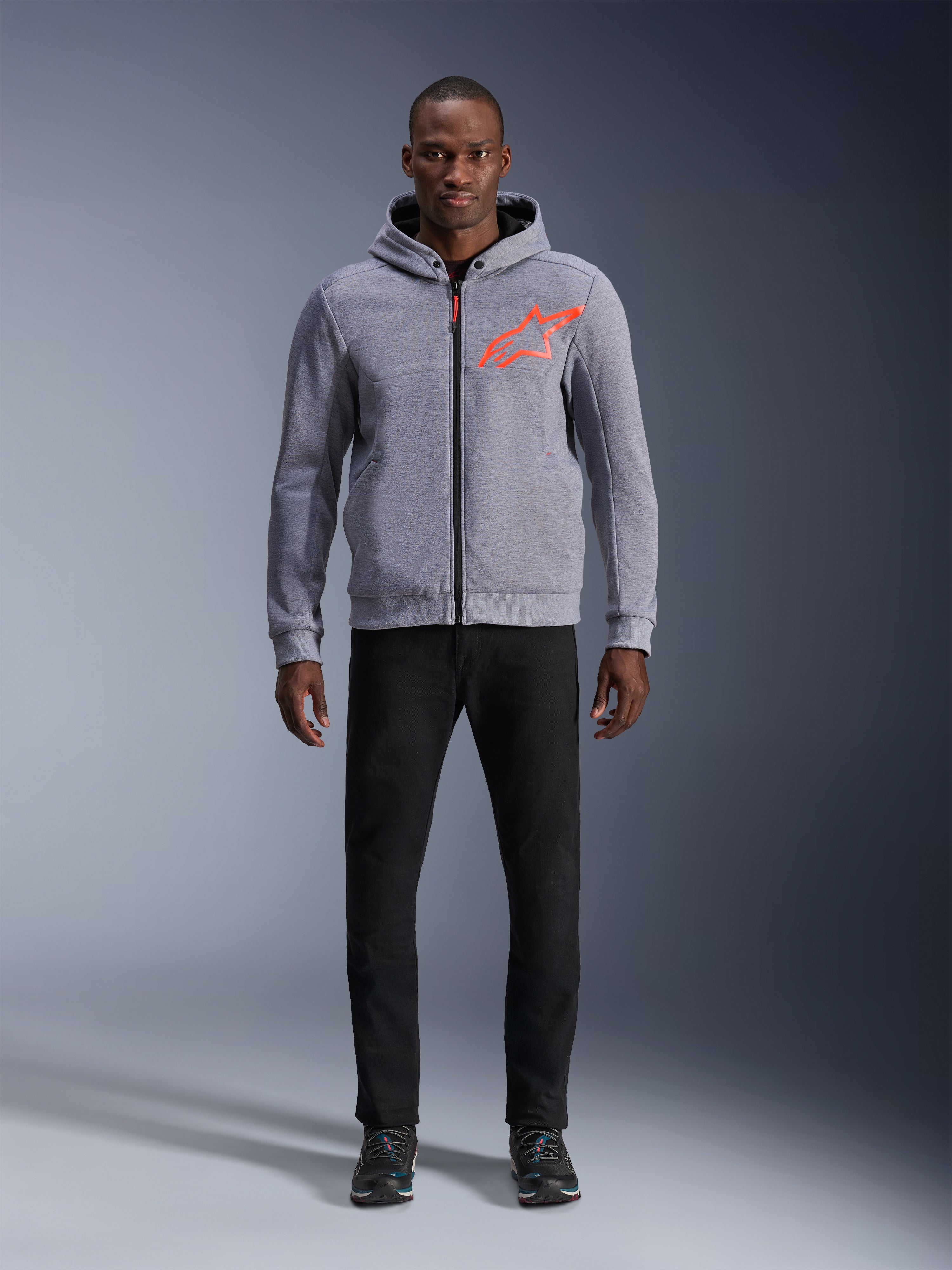 Alpinestars Chrome V2 Sport Hoodie in Gray Heather Red Fluo, lichtgrijze tech fleece hoodie met een fel oranjerood ster-logo op de borst, model draagt zwarte broek en donkere sneakers, casual stedelijke motorstijl