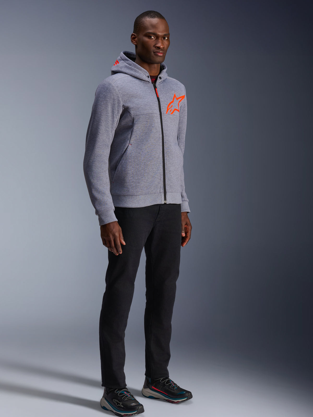 Alpinestars Chrome V2 Sport Hoodie, grijs gemêleerd en fluo rood, gedragen door een mannelijk model met zwarte broek en donkere sportschoenen, met een feloranje sterlogo op de borst en capuchon, casual stedelijke motorkleding