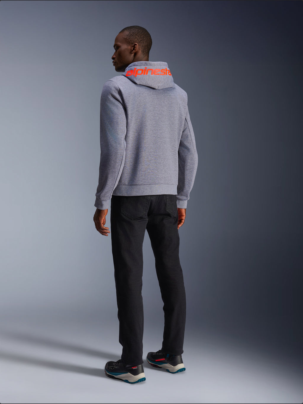 Alpinestars Chrome V2 Sport Hoodie in Gray Heather, achteraanzicht met een groot Red Fluo Alpinestars-woordmerk op de capuchon, gedragen door een model met een zwarte broek en donkere motorschoenen, casual stedelijke motorkleding