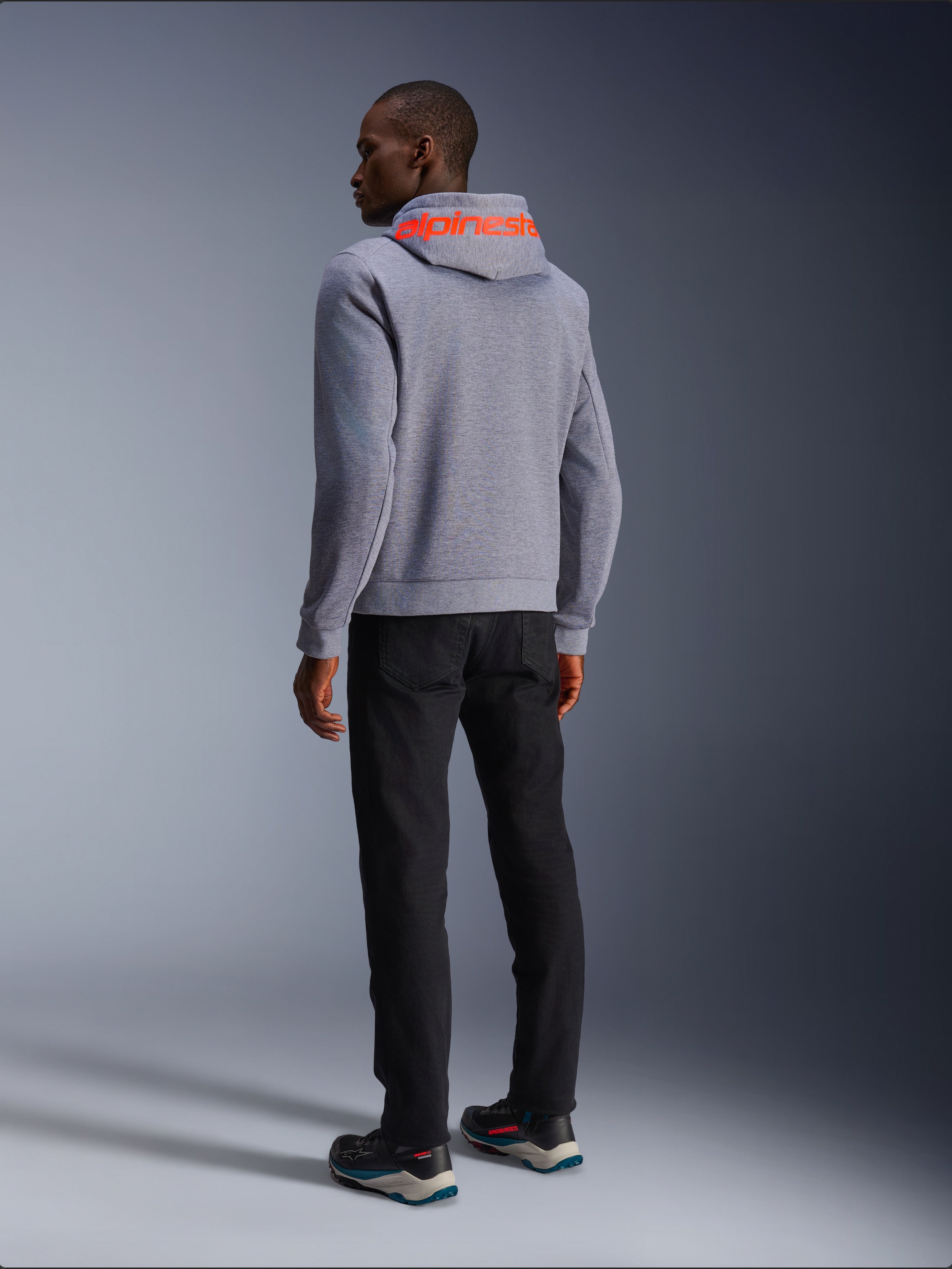 Alpinestars Chrome V2 Sport Hoodie in Gray Heather, achteraanzicht met een groot Red Fluo Alpinestars-woordmerk op de capuchon, gedragen door een model met een zwarte broek en donkere motorschoenen, casual stedelijke motorkleding