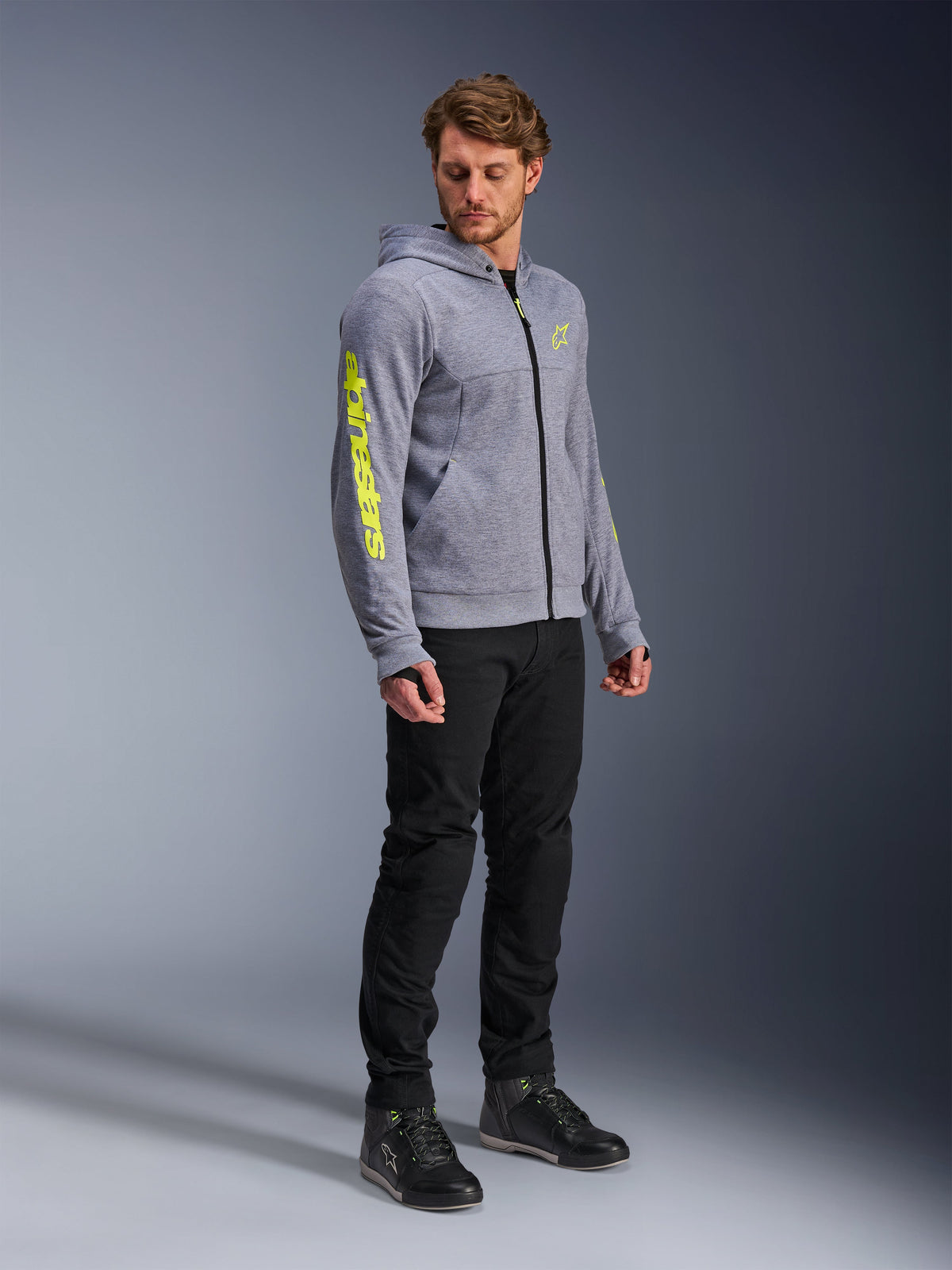 Alpinestars Chrome V2 Sport Hoodie, grijs gemêleerd en fluogeel, gedragen door een mannelijk model met zwarte broek en zwarte motorschoenen, met opvallende gele branding op de mouw en borst, casual stedelijke motorkleding