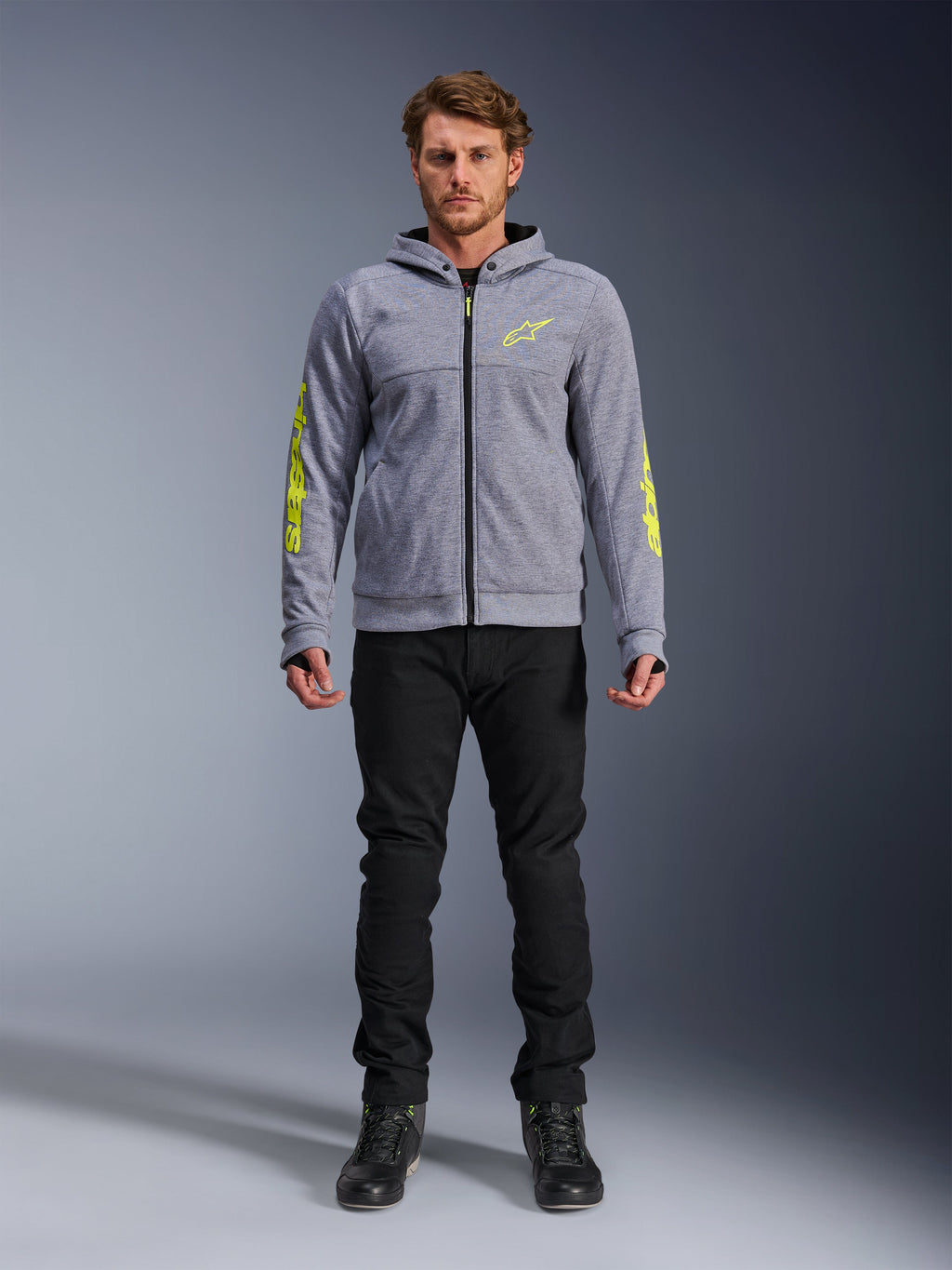 Alpinestars Chrome V2 Sport Hoodie, grijs gemêleerd met fluorescerend gele accenten, stedelijke motorkleding, getoond met zwarte broek en zwarte motorschoenen, technische fleece met opvallende gele branding op de mouwen en borst, casual straatstijl voor tijdens het rijden