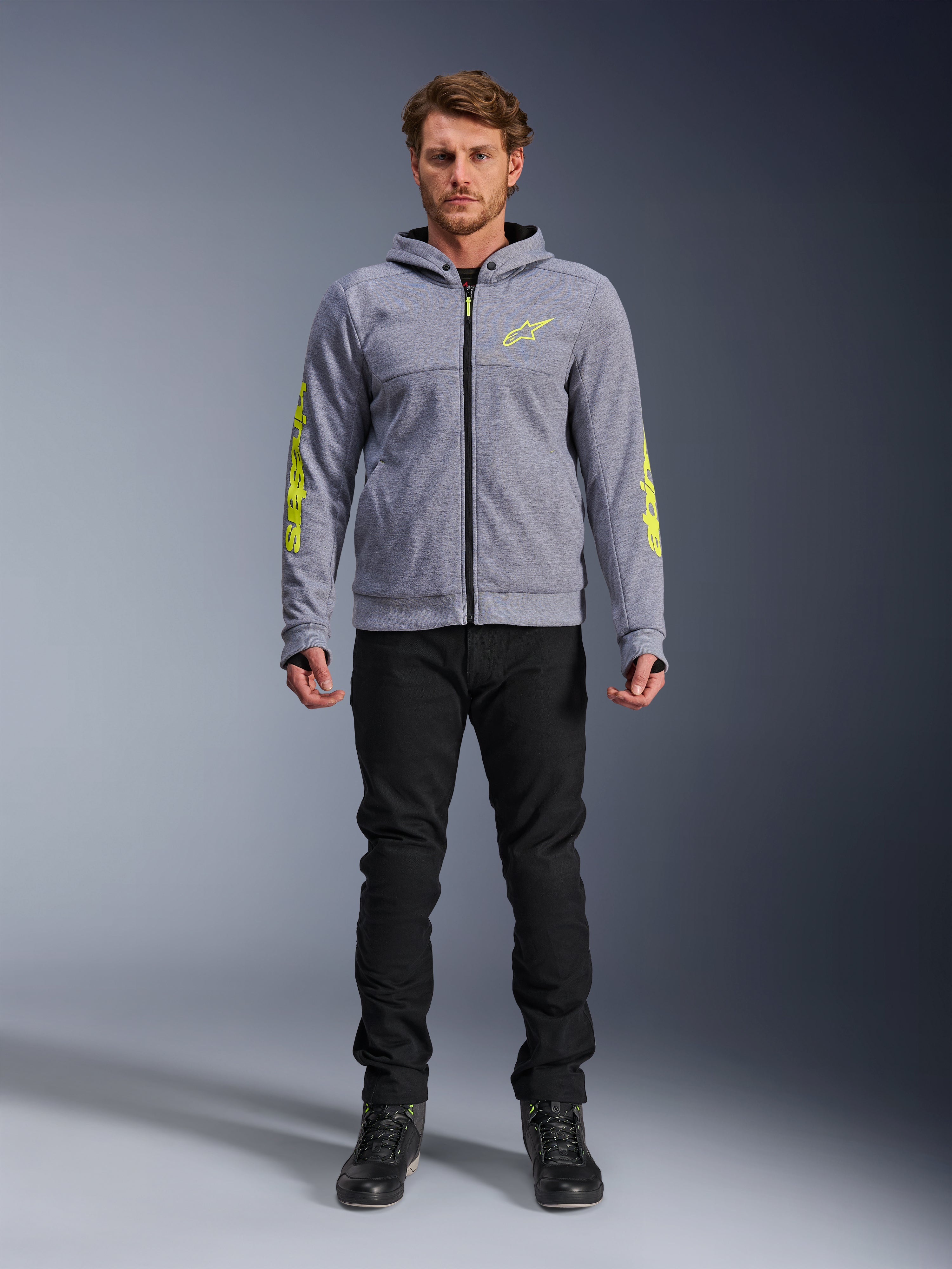 Alpinestars Chrome V2 Sport Hoodie, grijs gemêleerd met fluorescerend gele accenten, stedelijke motorkleding, getoond met zwarte broek en zwarte motorschoenen, technische fleece met opvallende gele branding op de mouwen en borst, casual straatstijl voor tijdens het rijden