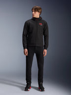 Alpinestars MM93 Radium Tech Hoodie, zwarte urban motorhoodie, voorzien van een rood 93-logo en wit sterlogo op de borst, gedragen door een model met een zwarte broek en bijpassende motorschoenen, slanke winddichte technische shell voor ritten in de stad.