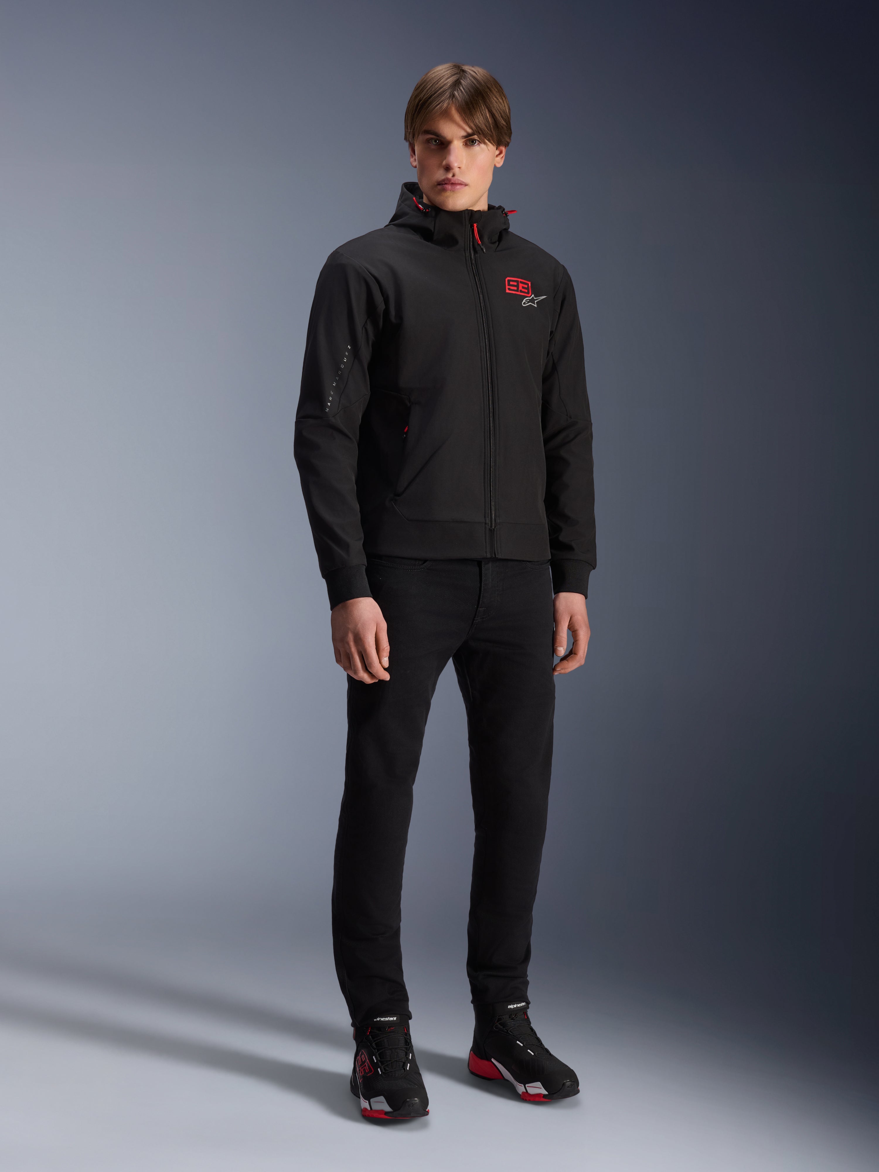 Alpinestars MM93 Radium Tech Hoodie, urban motorjas, zwart met felrode accenten, gedragen door model met zwarte broek en motorschoenen, voorzien van rood '93' logo op de borst en Marc Marquez branding op de mouw, casual technische streetwear voor stedelijk rijden