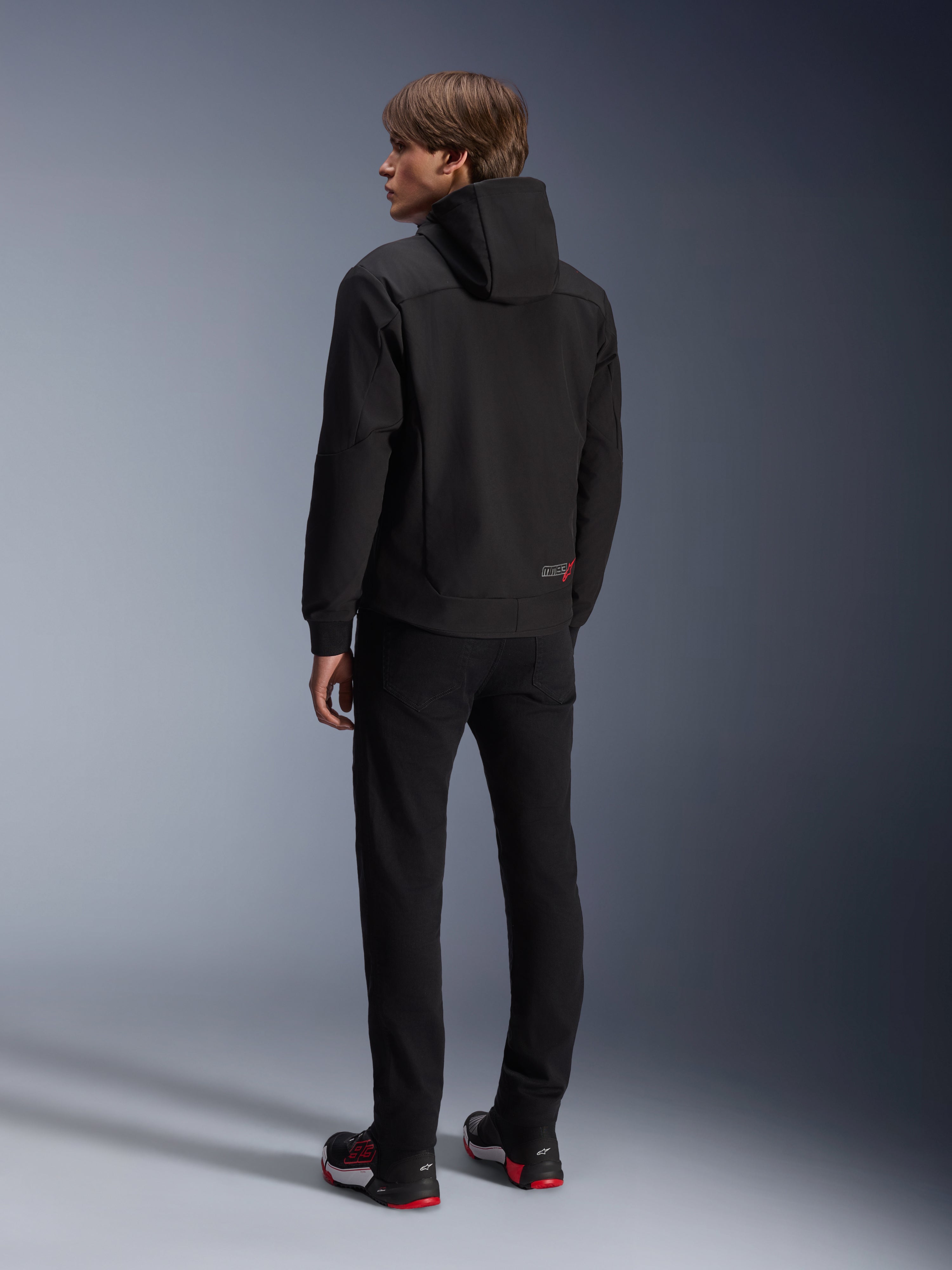 Alpinestars MM93 Radium Tech Hoodie, zwart, urban motorhoodie achteraanzicht, met een klein rood en wit MM93-logo op de onderste zoom, atletische pasvorm met capuchon, gedragen met een zwarte broek en bijpassende rood-zwarte sneakers