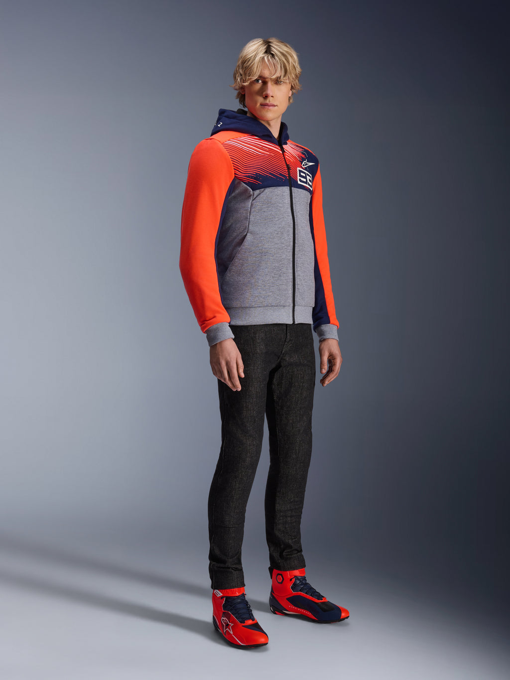 Alpinestars MM93 Chrome V2 Sport Hoodie, urban motorhoodie, grijs gemêleerde body met donkerblauwe borst en felrode fluo mouwen, voorzien van het 93-logo op de borst, gedragen door een model met slim-fit zwarte broek en bijpassende rode en blauwe hoge motorschoenen, moderne atletische pasvorm voor rijden op de weg
