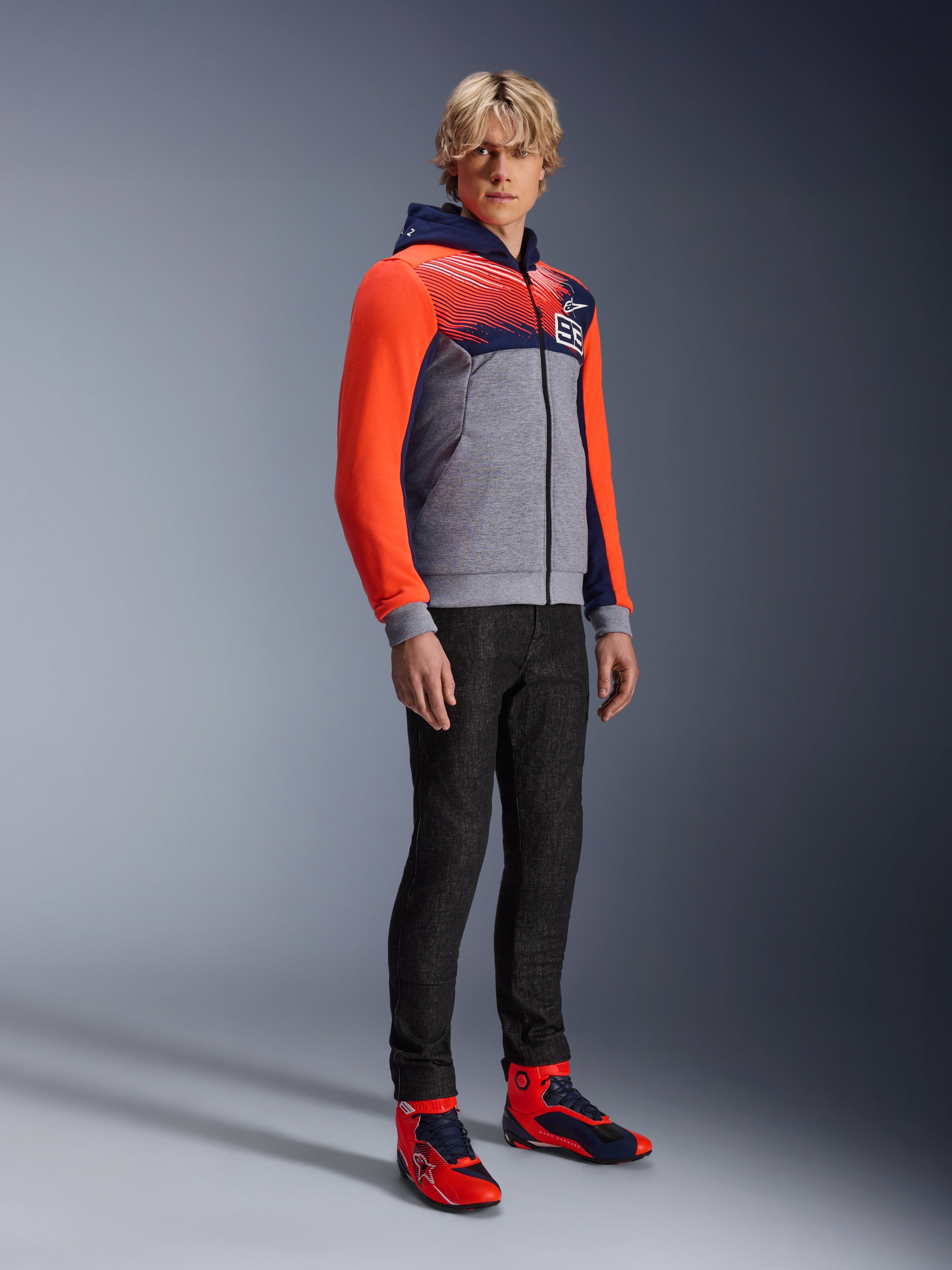 Alpinestars MM93 Chrome V2 Sport Hoodie, urban motorhoodie, grijs gemêleerde body met donkerblauwe borst en felrode fluo mouwen, voorzien van het 93-logo op de borst, gedragen door een model met slim-fit zwarte broek en bijpassende rode en blauwe hoge motorschoenen, moderne atletische pasvorm voor rijden op de weg
