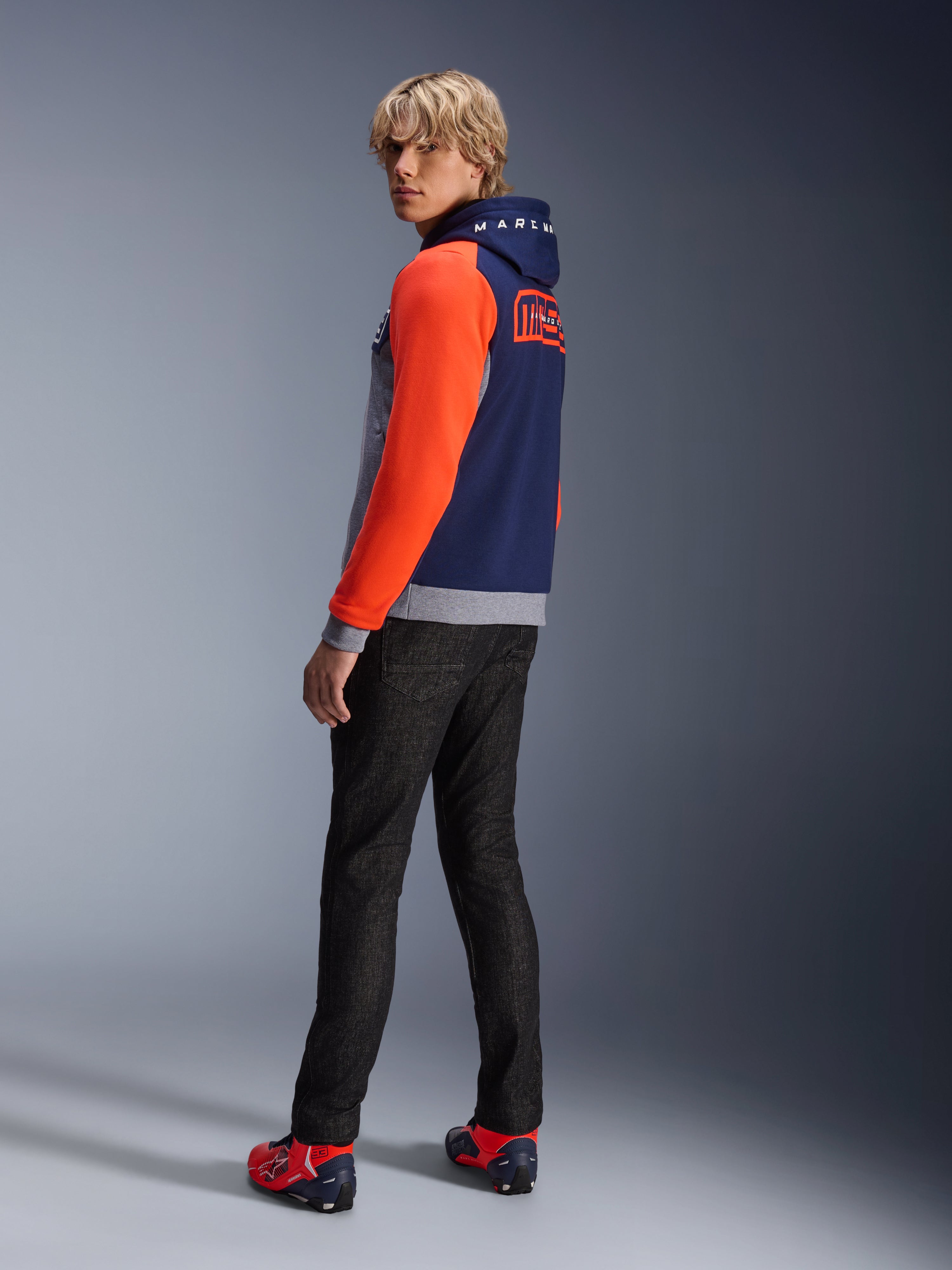 Alpinestars MM93 Chrome V2 Sport Hoodie, Blue Navy Gray Mel Red Fluo, model draagt hoodie met felrode mouwen en MM93 Marc Marquez branding op de rug, gecombineerd met een donkere jeans en rode motorschoenen, casual stedelijke motorkleding