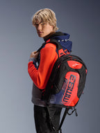 Mannelijk model draagt de Alpinestars MM93 Chrome V2 Sport Hoodie in Blue Navy Gray Mel en Red Fluo, gecombineerd met een bijpassende MM93 rugzak, casual stedelijke motorkleding met een modern atletisch silhouet.