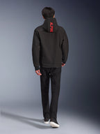 Alpinestars Radium Tech Hoodie, urban motorjas, zwart, achteraanzicht met felrode 'ALPINE'-tekst op de capuchon, gedragen met zwarte broek en sneakers, strakke technische shell voor woon-werkverkeer.