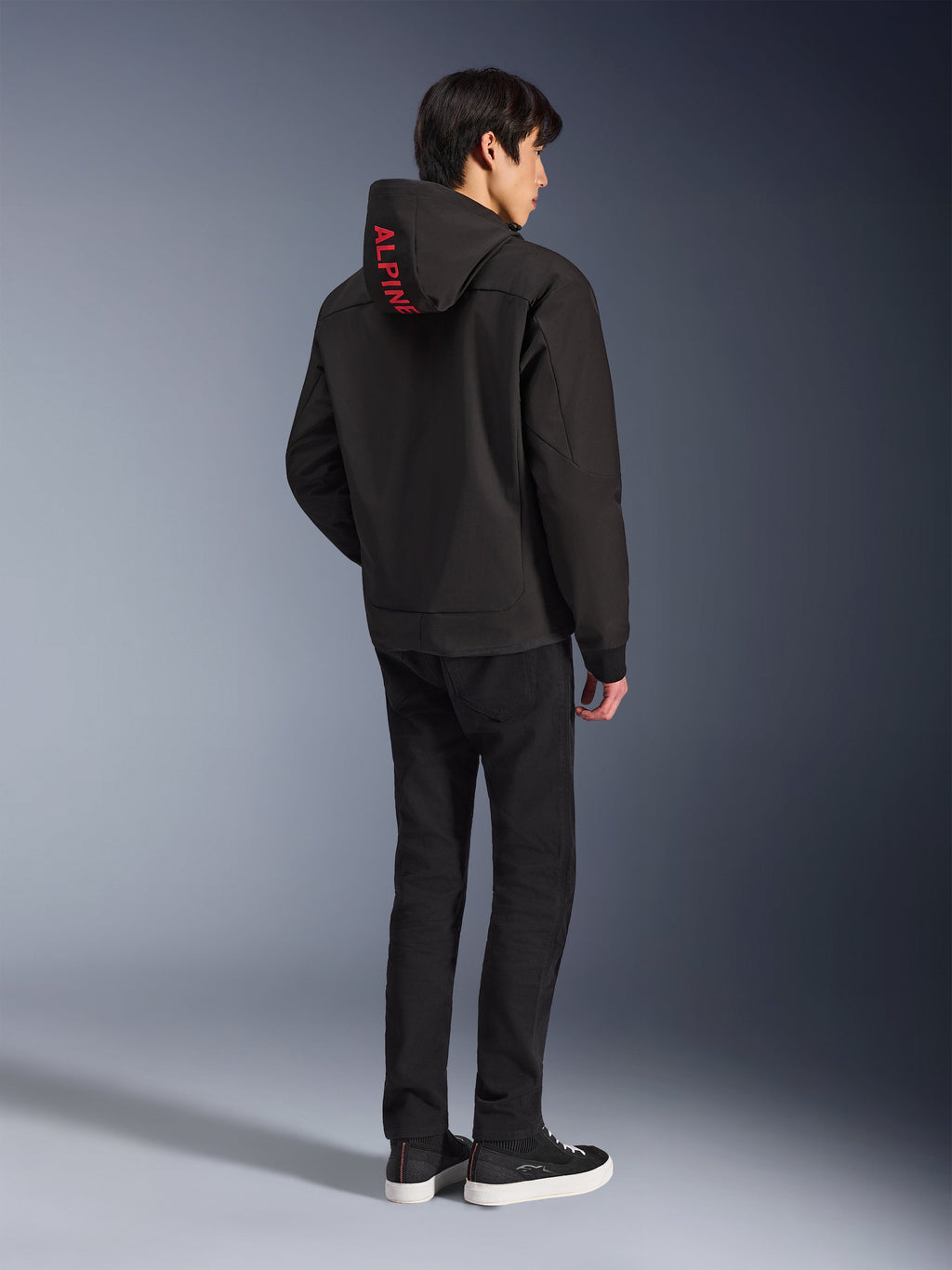 Alpinestars Radium Tech Hoodie, zwart met rode Alpinestars-branding op de capuchon, achteraanzicht op een mannelijk model, gedragen met zwarte broek en sneakers, technische stedelijke motorkleding