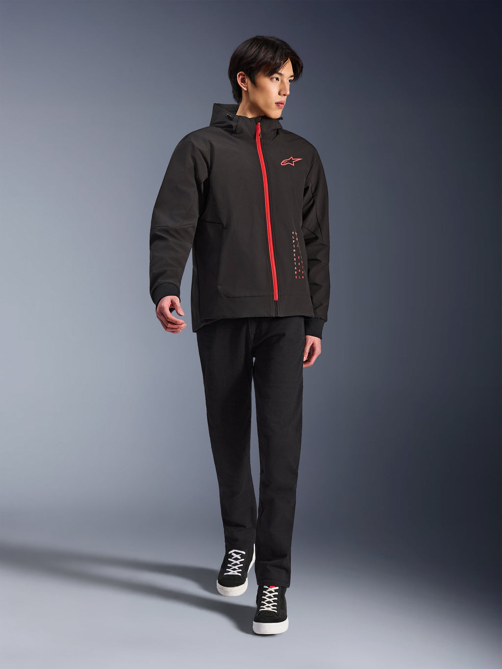 Alpinestars Radium Tech Hoodie in Black Bright Red, stedelijke motorjas gedragen door een model, voorzien van een rode ritssluiting aan de voorzijde en een logo op de borst, gecombineerd met een zwarte broek en sneakers voor een moderne woon-werk uitstraling.