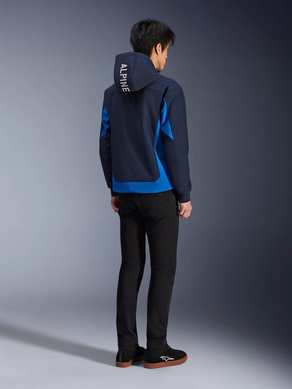 Alpinestars Radium Tech Hoodie, Night Navy en Royal Blue, achteraanzicht van model in de technische shell hoodie met witte 'ALPINE' tekst op de capuchon, gecombineerd met een zwarte broek en schoenen, stedelijke motoruitrusting