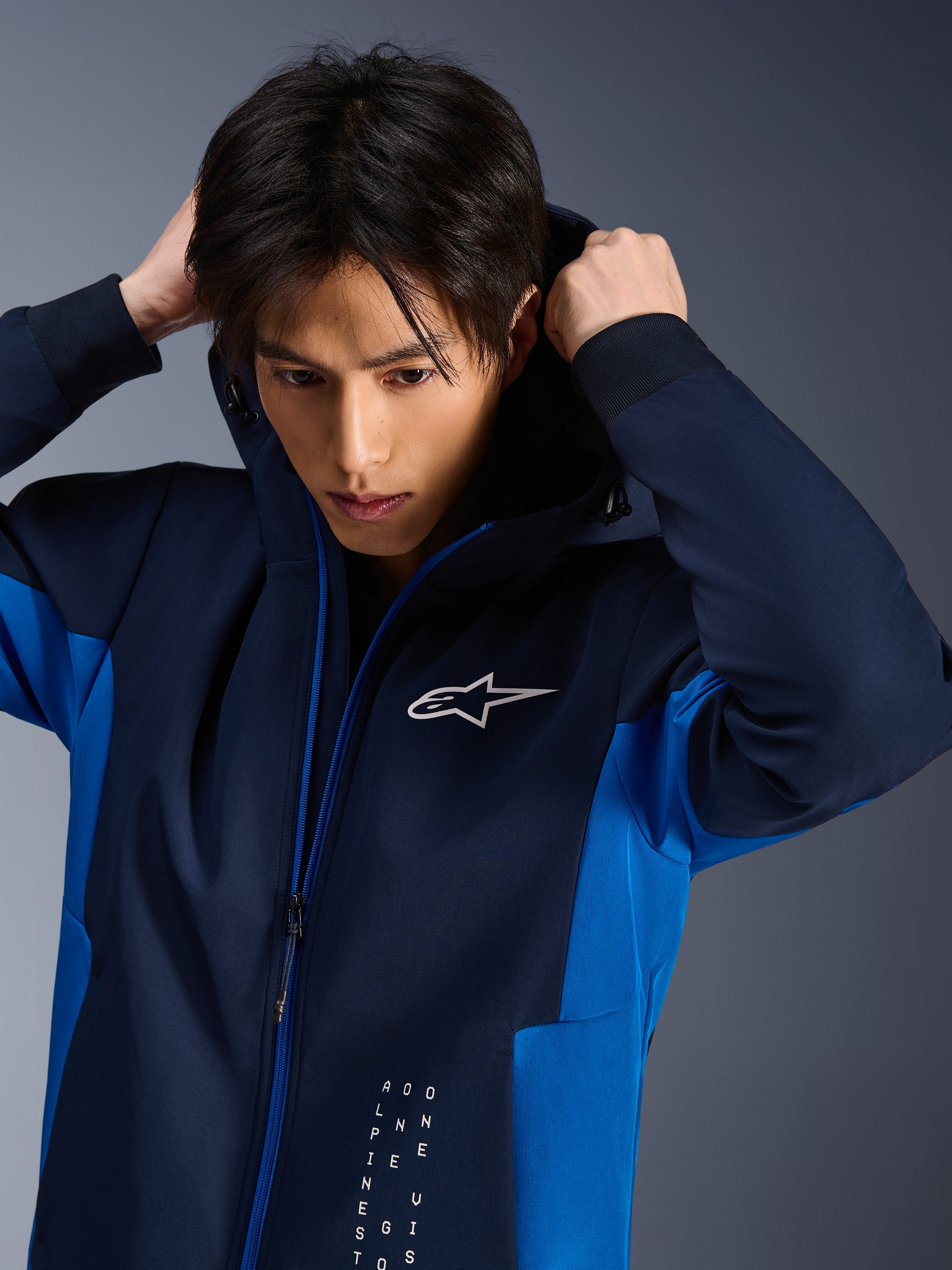 Alpinestars Radium Tech Hoodie, stedelijke motorjas, Night Navy en Royal Blue, gedragen door een model dat de capuchon verstelt, met een wit Astars-logo op de borst en een technisch 3L shell-ontwerp met atletische pasvorm.
