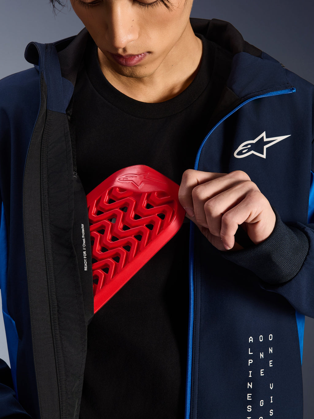 Alpinestars Radium Tech Hoodie in Night Navy en Royal Blue, close-up van een rode Nucleon borstprotector die in de binnenzak wordt geplaatst, voorzien van 'READY FOR // Chest Protector' tekst op de voering en een wit sterlogo op de borst.