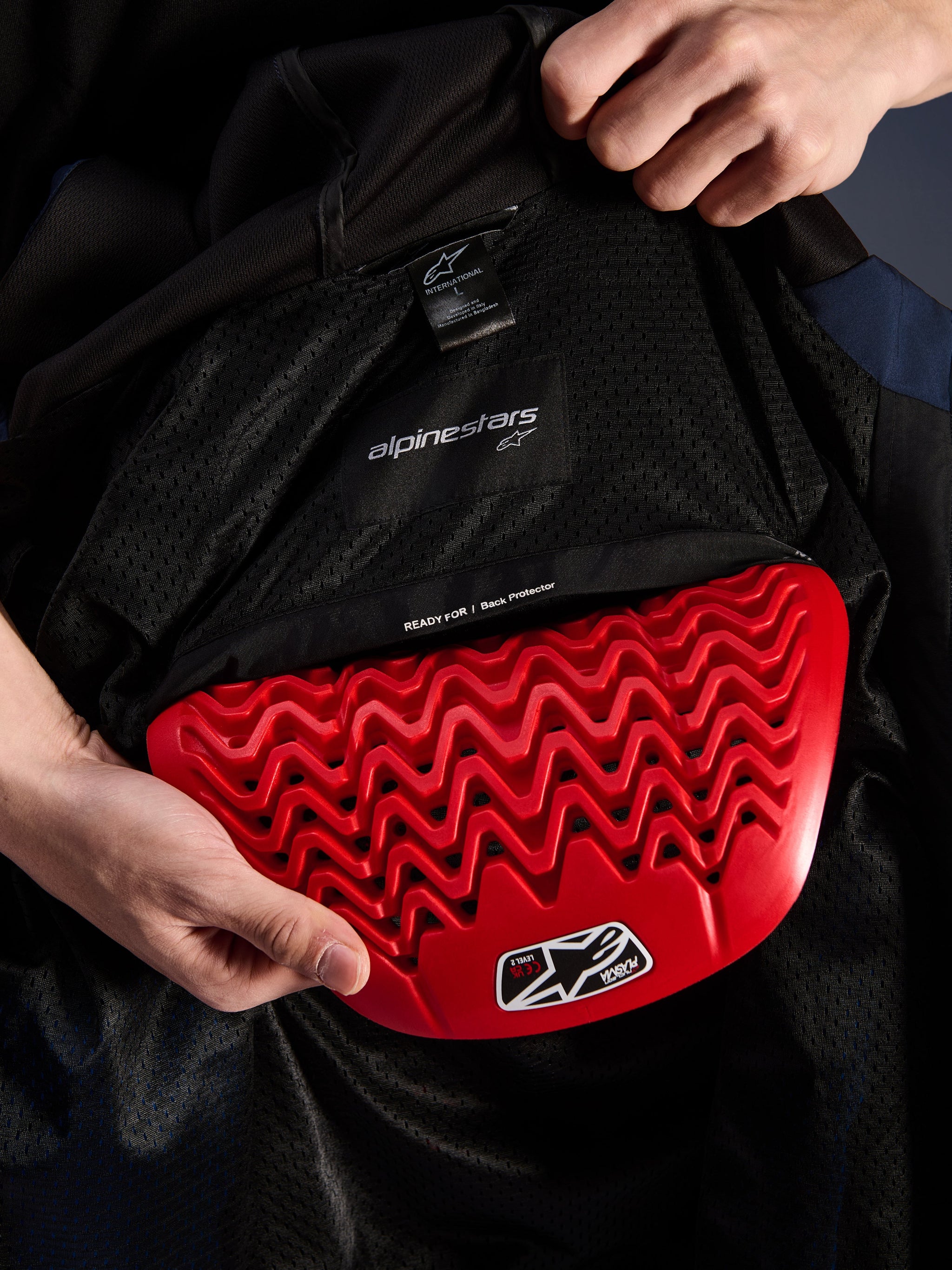 Alpinestars Radium Tech Hoodie interior detail, red Nucleon Plasma back protector being inserted into the back protector pocket, black mesh lining with 'READY FOR | Back Protector' text, wavy honeycomb armor design, technical urban motorcycle jacket protection features