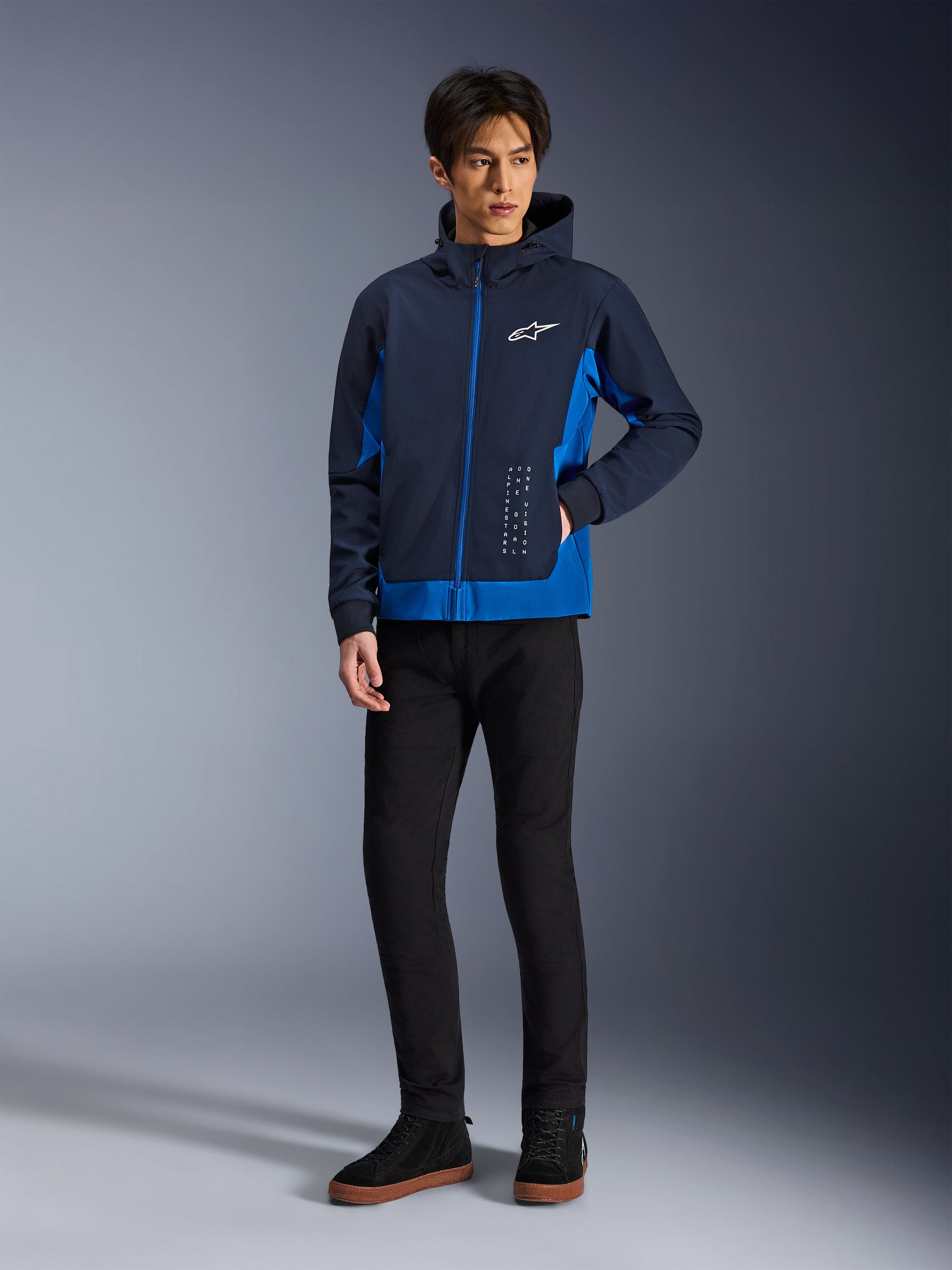 Alpinestars Radium Tech Hoodie, Urban Motorcycle Jacket, Night Navy and Royal Blue, worn with black pants and black sneakers, featuring a white Alpinestars logo on the chest and vertical text detailing, casual athletic fit for city riding