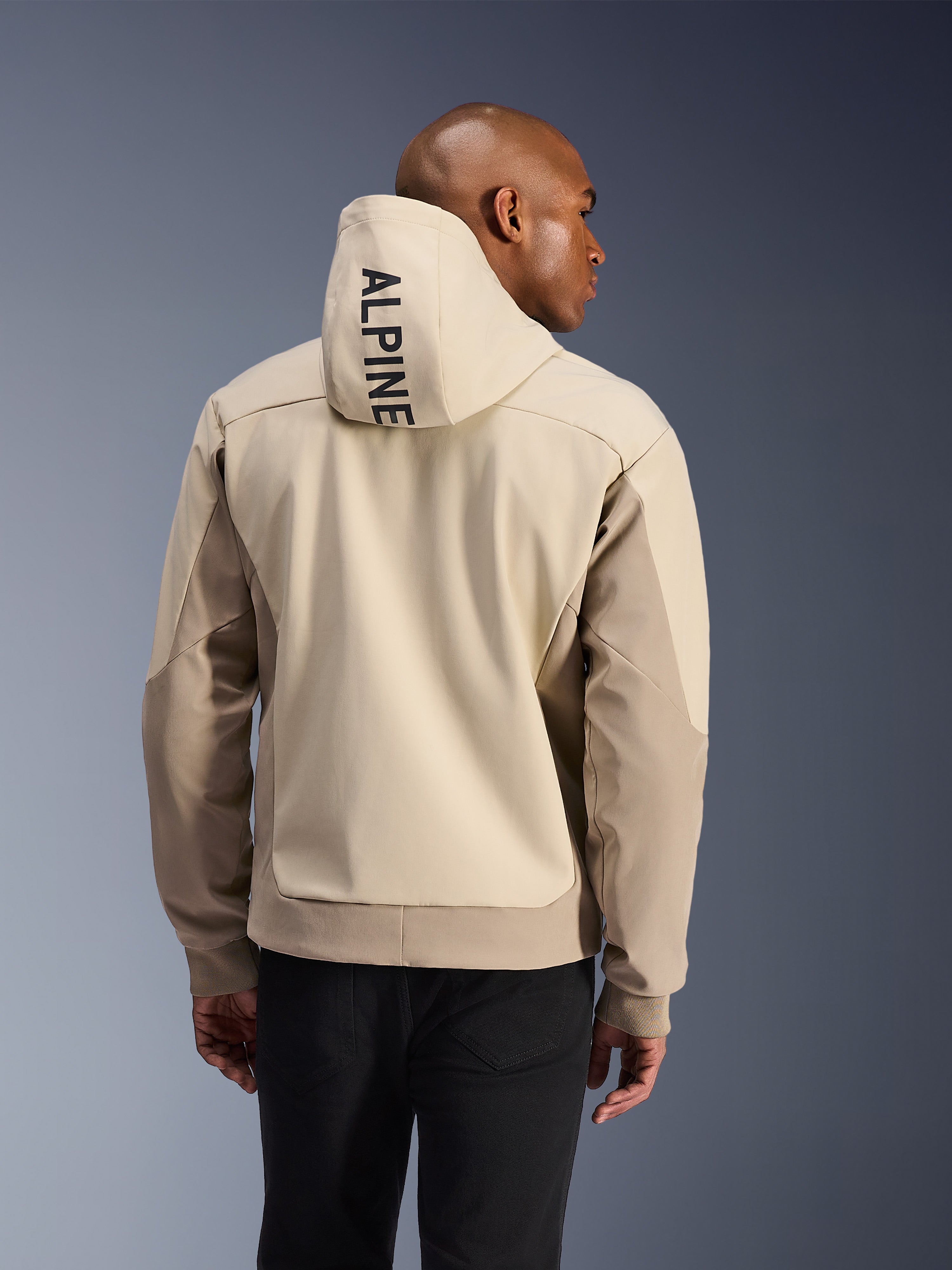 Alpinestars Radium Tech Hoodie, achteraanzicht van een man die de Mastic Aluminum technische hoodie draagt, met verticale ALPINE-tekst op de capuchon, lichtbeige en zandkleurige panelen, geribde manchetten, stedelijke motorjas