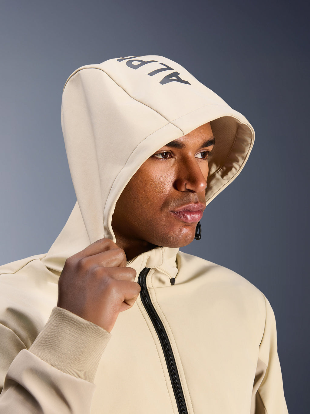 Alpinestars Radium Tech Hoodie in Mastic Aluminum, close-up van een man met de capuchon op, voorzien van Alpinestars-branding op de bovenkant, stedelijke motorjas met technische winddichte shell, moderne streetwear-esthetiek voor stadsritten