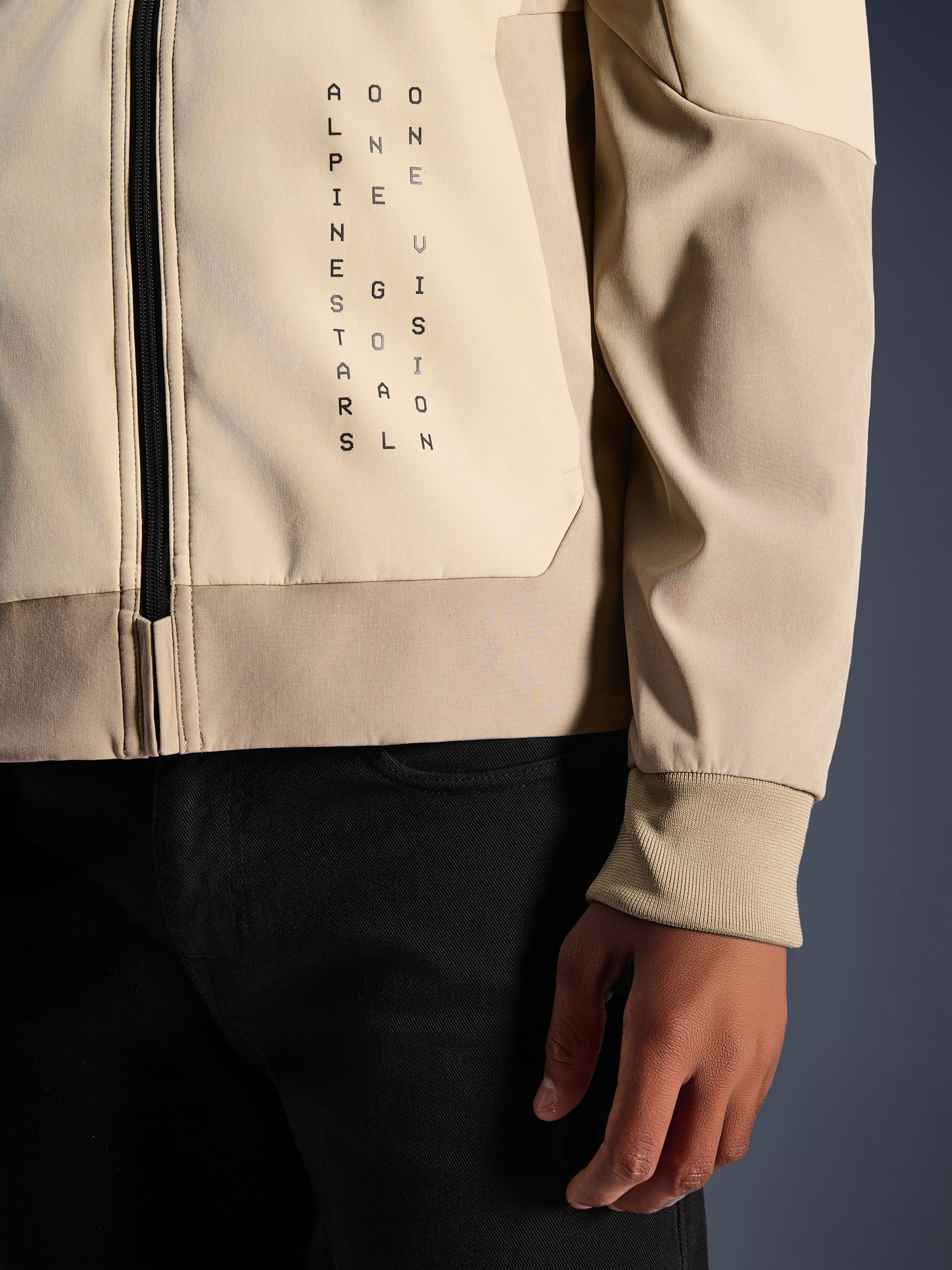 Alpinestars Radium Tech Hoodie in Mastic Aluminum, close-up van de borst en mouw, verticale zwarte tekstbranding 'Alpinestars One Goal One Vision', geribde stretch manchet en ritsdetail, urban motorjas van textiel.