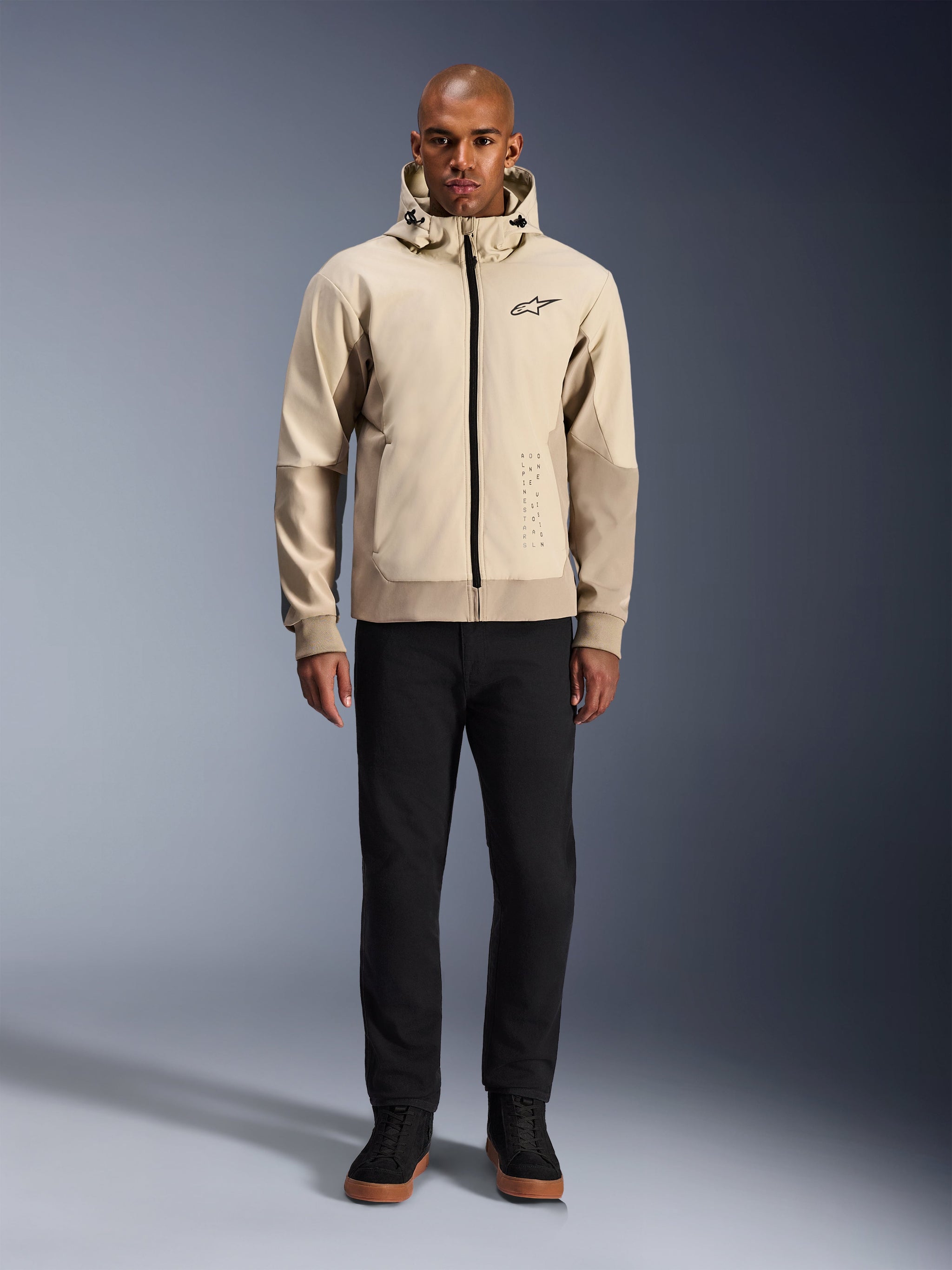 Alpinestars Radium Tech Hoodie in Mastic Aluminum, stedelijke motorhoodie gedragen door een mannelijk model met zwarte broek en zwarte laarzen, voorzien van een zwarte ritssluiting over de volledige lengte, klein logo op de borst, atletische pasvorm voor stadsritten