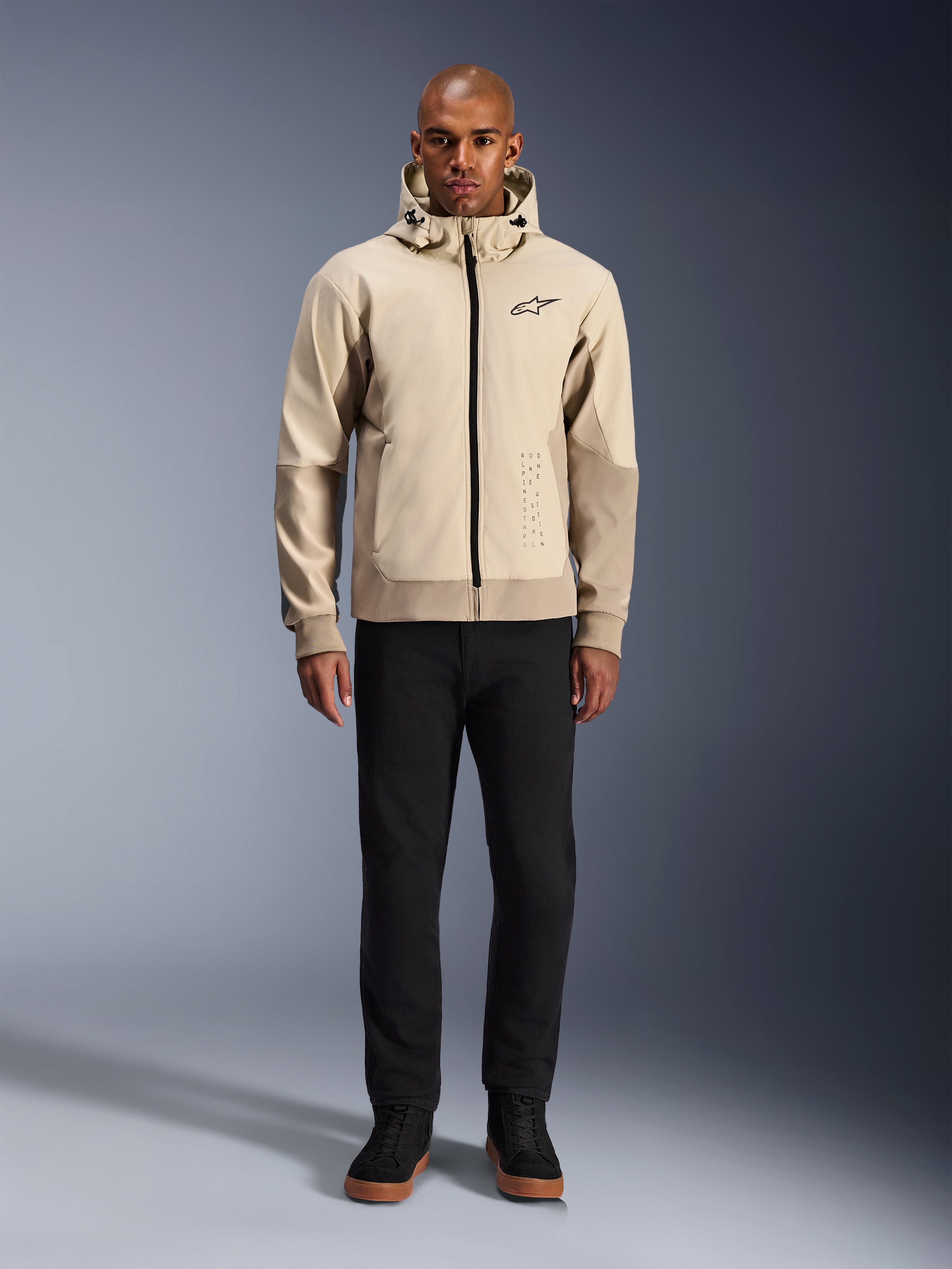Alpinestars Radium Tech Hoodie in Mastic Aluminum, stedelijke motorhoodie gedragen door een mannelijk model met zwarte broek en zwarte laarzen, voorzien van een zwarte ritssluiting over de volledige lengte, klein logo op de borst, atletische pasvorm voor stadsritten