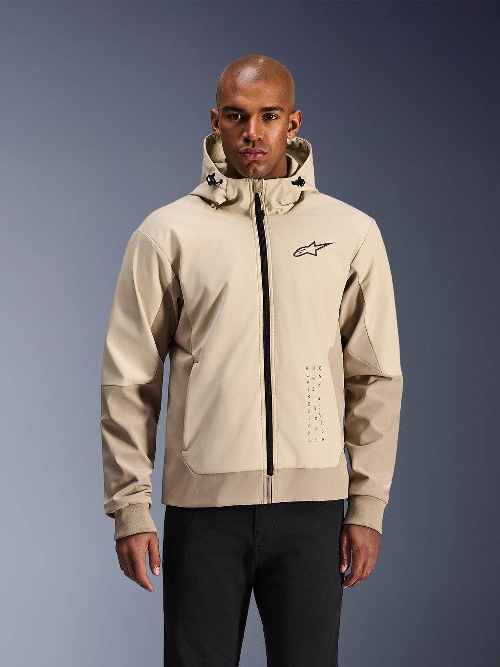 Alpinestars Radium Tech Hoodie in Mastic Aluminum, lichtbeige en tan stedelijke motorjas, zwarte rits aan de voorzijde en klein borstlogo, gedragen door een man in een zwarte broek, technische winddichte shell met eigentijdse streetwear-stijl